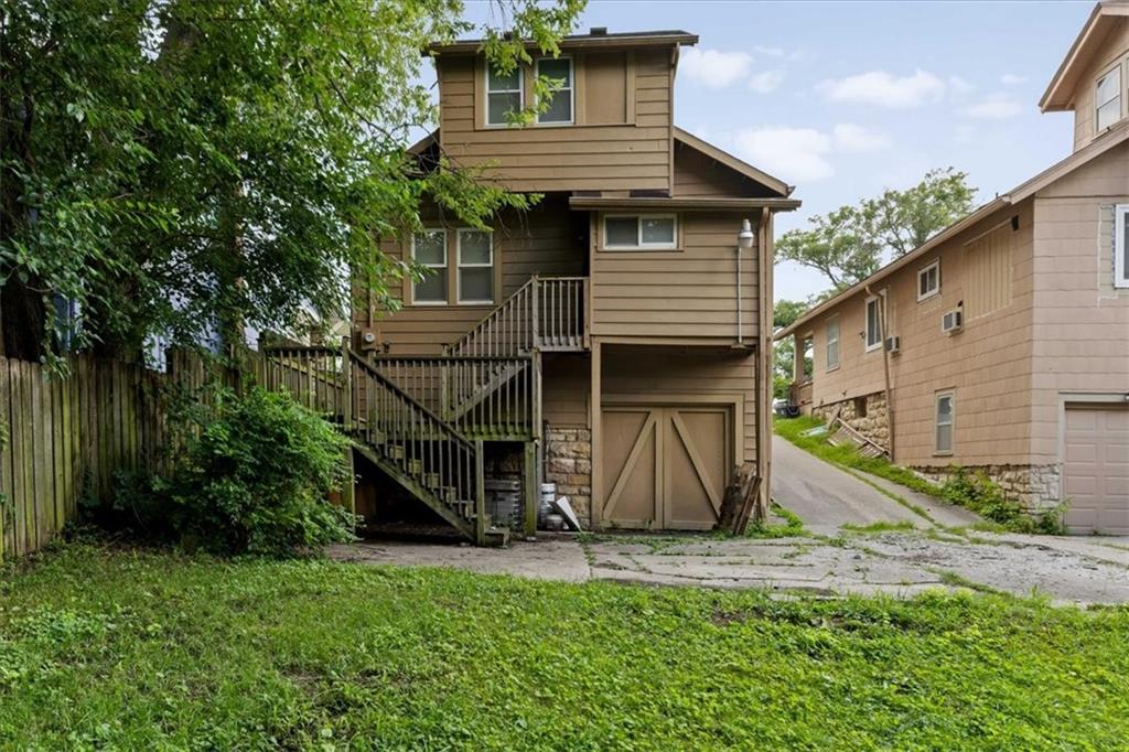 Rear of a tan home with an elevated wooden staircase, deck, and garage doors beneath the house.