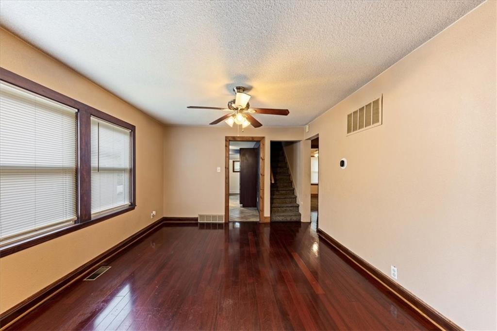 Empty living room with dark hardwood floors, wide windows, and a staircase at the back.