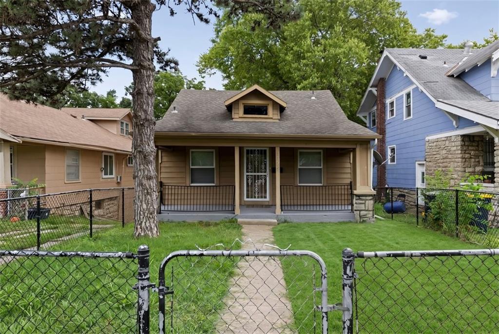 Tan single-story house with a covered porch and dormer behind a chain-link fence, shaded by a large tree.