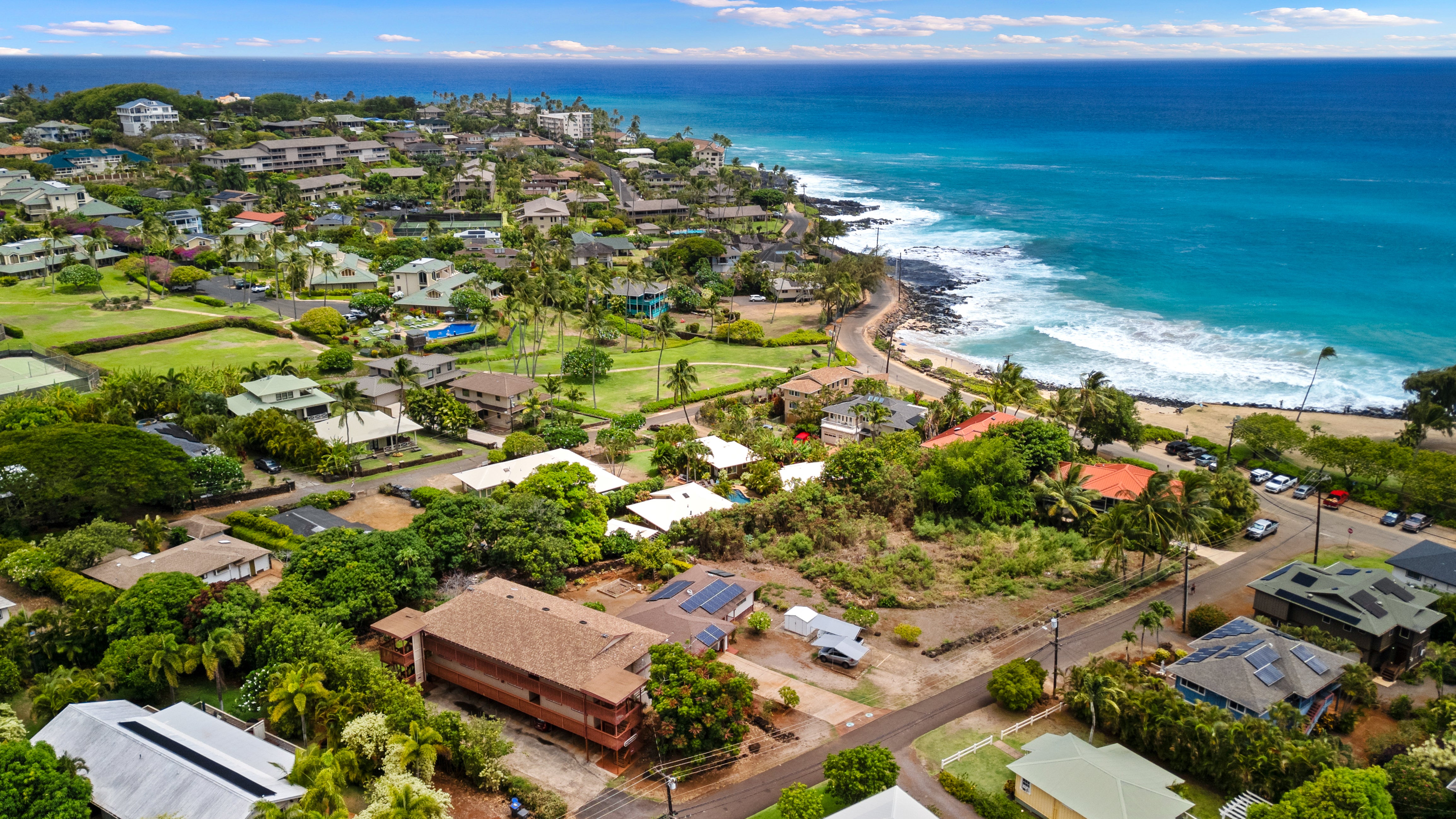 Aerial view of coastal homes and tropical greenery beside a blue ocean and rocky shoreline.