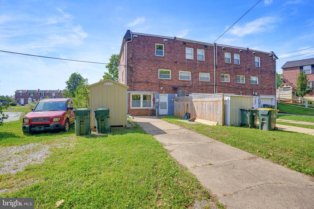 Rear of a brick townhome with a paved walkway, lawn, utility sheds, and trash bins.