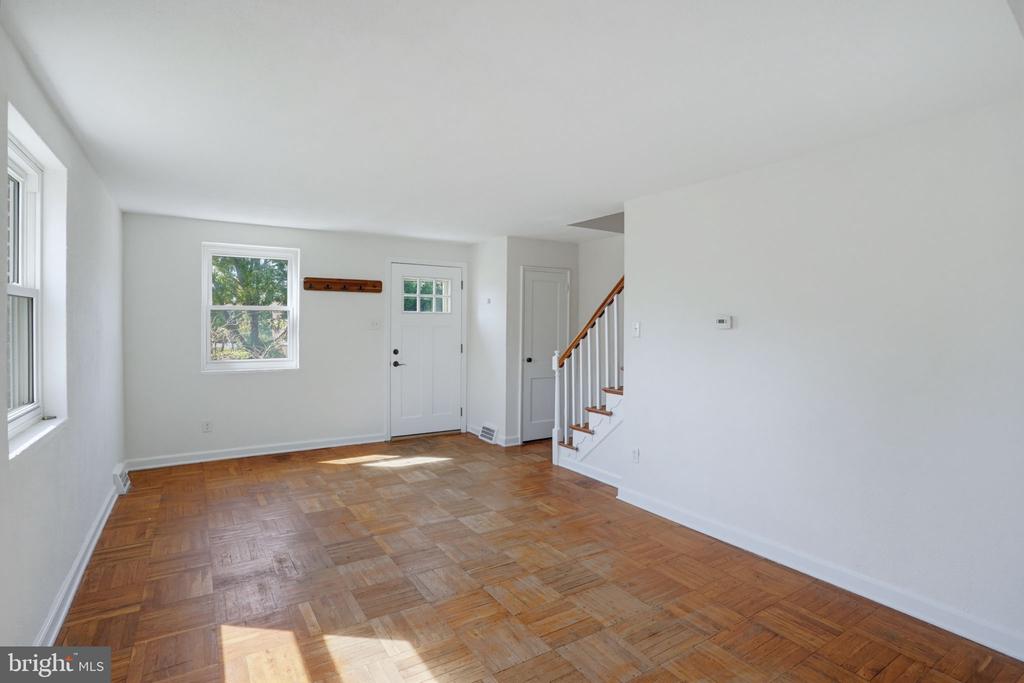 Vacant living room with parquet flooring, an entry door, and stairs beside a large white wall.