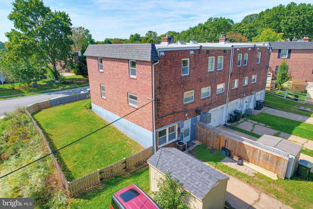 Aerial view of a red-brick townhome building and fenced backyards with sheds and a parked vehicle.