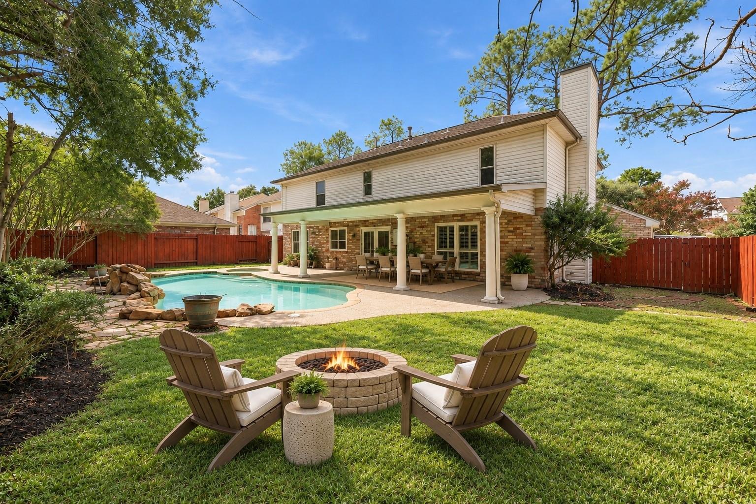 Backyard pool and covered patio behind a two-story home, with a stone fire pit and Adirondack chairs.