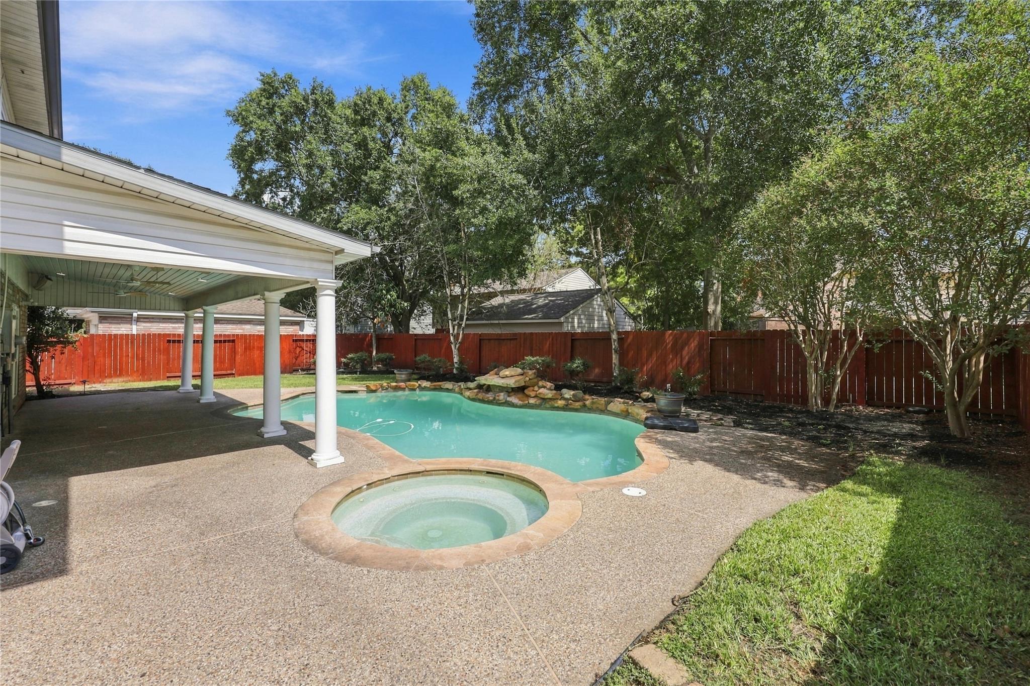 Backyard pool and circular spa beside a covered patio, with mature trees and a red privacy fence.