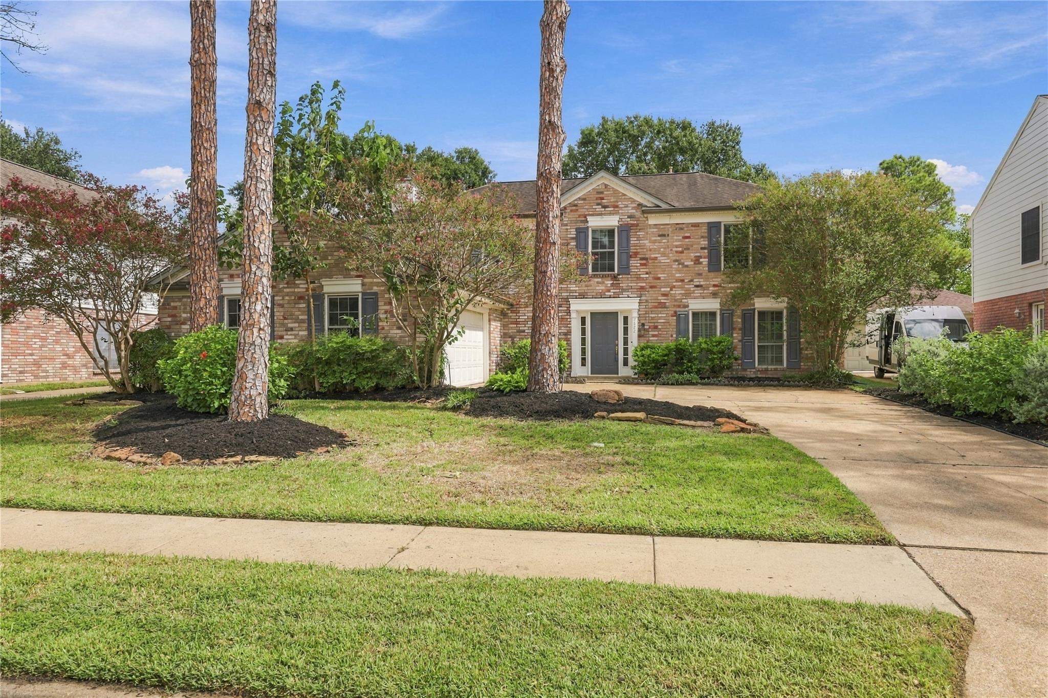 Two-story brick house with a garage, wide driveway, landscaped lawn, and tall pine trees.