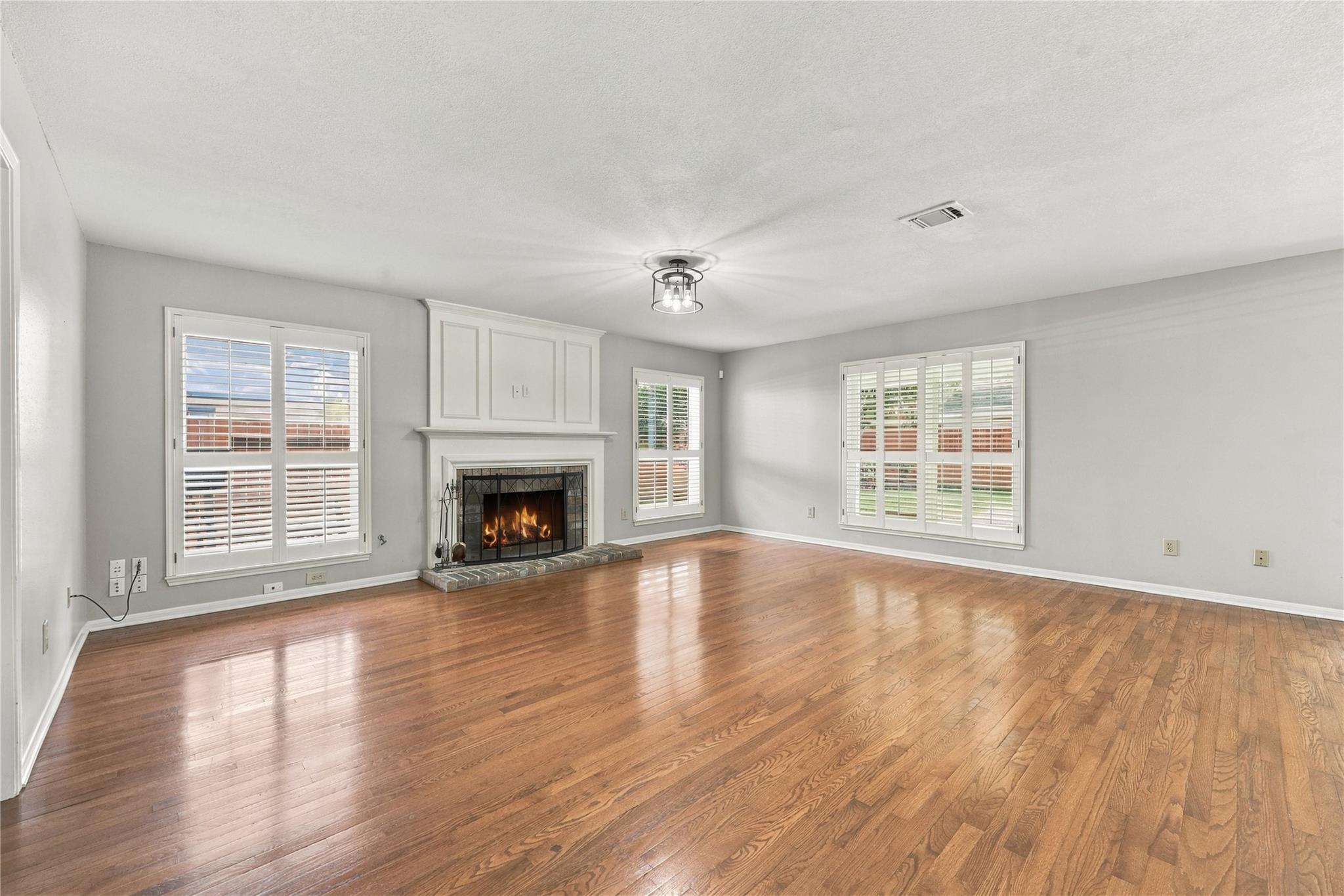Empty living room with hardwood floors, a white fireplace, and shuttered windows.