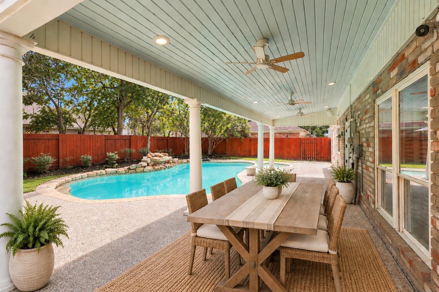 Covered patio dining area beside a curved swimming pool, with wicker chairs and a red privacy fence.