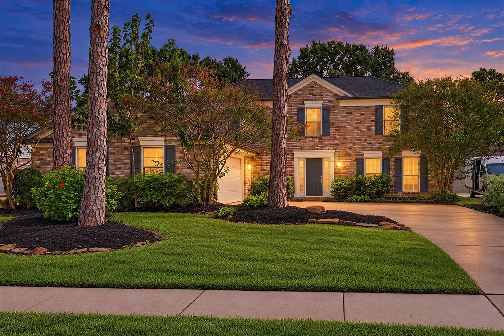Two-story brick house at dusk, framed by tall trees with warmly lit windows and a landscaped lawn.