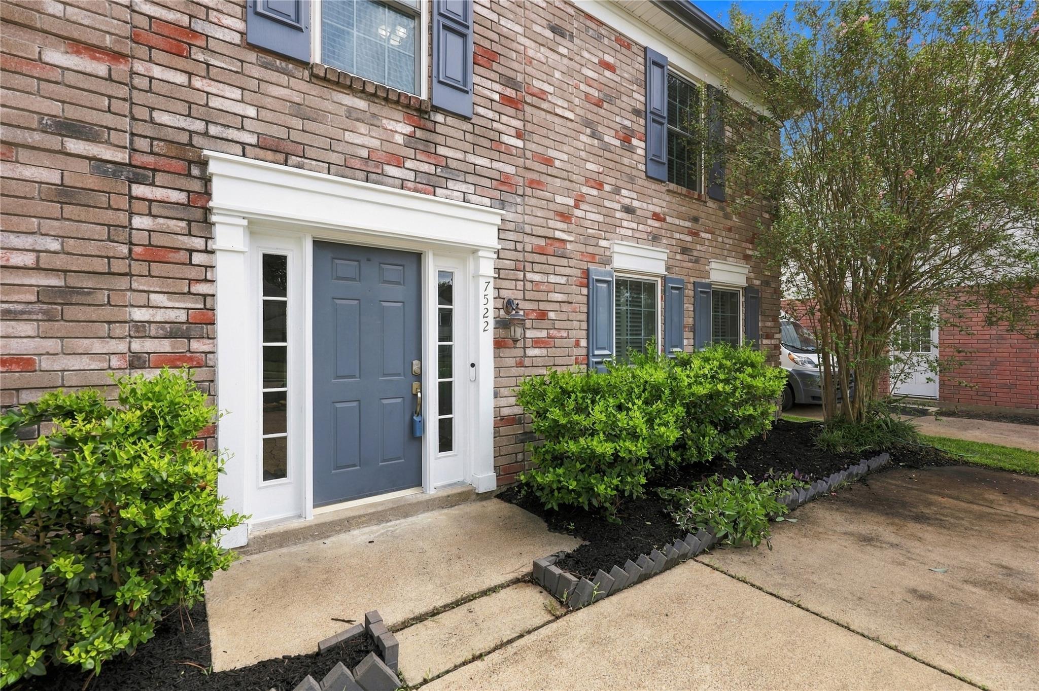 Brick home entrance with a blue front door, white trim, and landscaped shrubs beside the walkway.