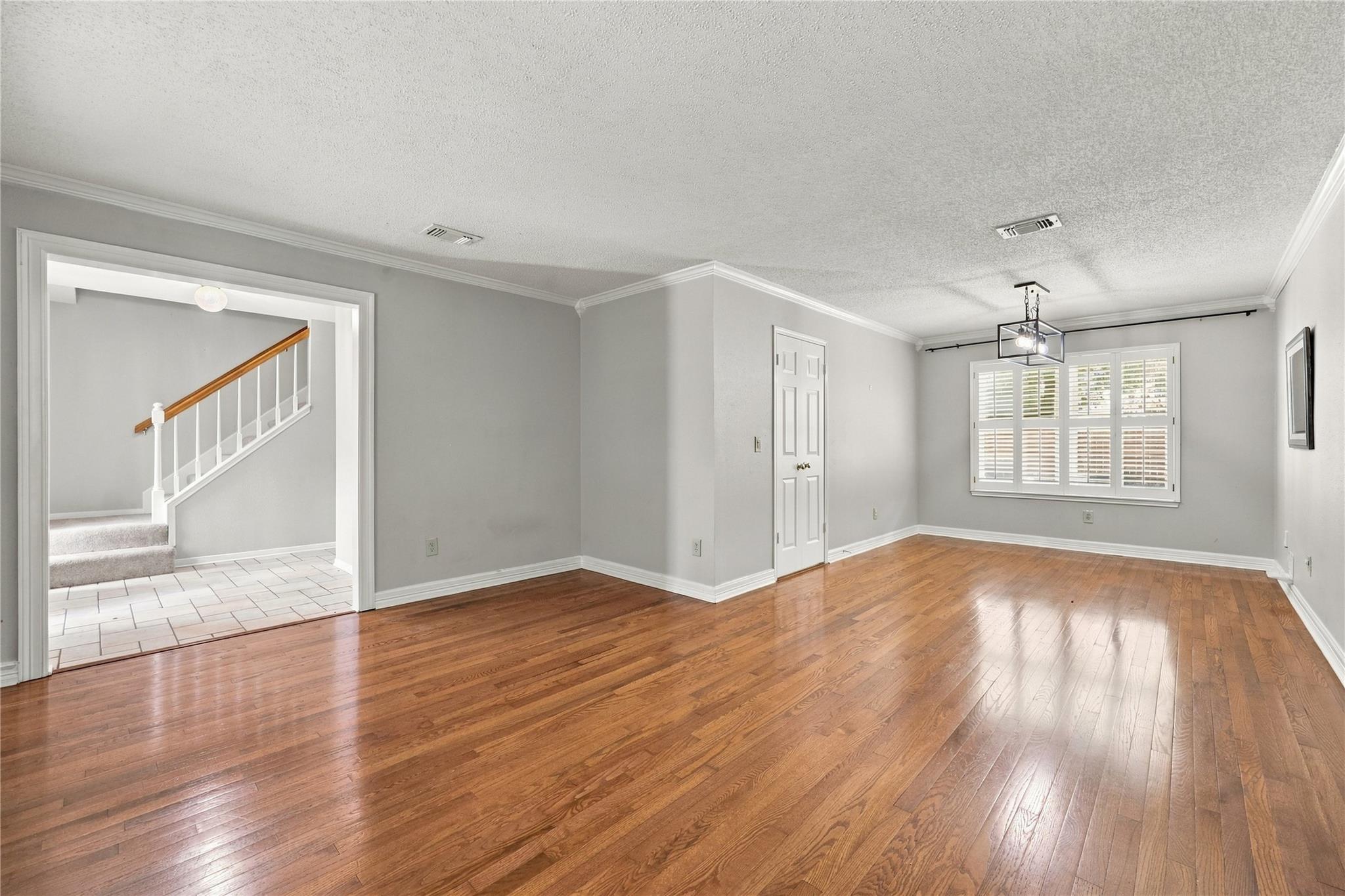 Empty room with polished hardwood floors, plantation shutters, and a view of the staircase through an open doorway.