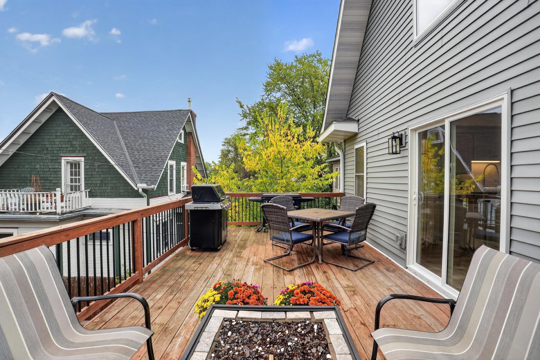 Wooden deck with outdoor dining set, grill, and fire feature overlooking autumn trees.