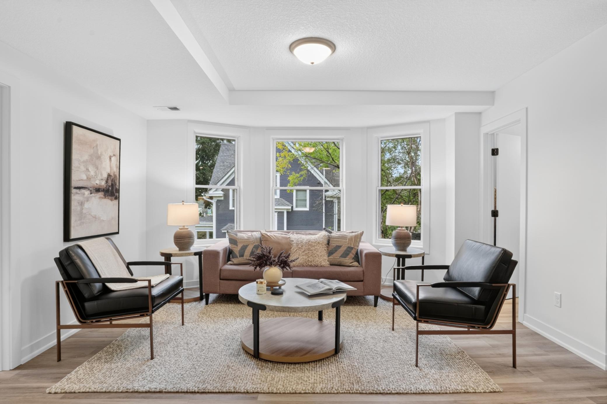 Staged living room with a mauve sofa, black leather armchairs, and three large windows.