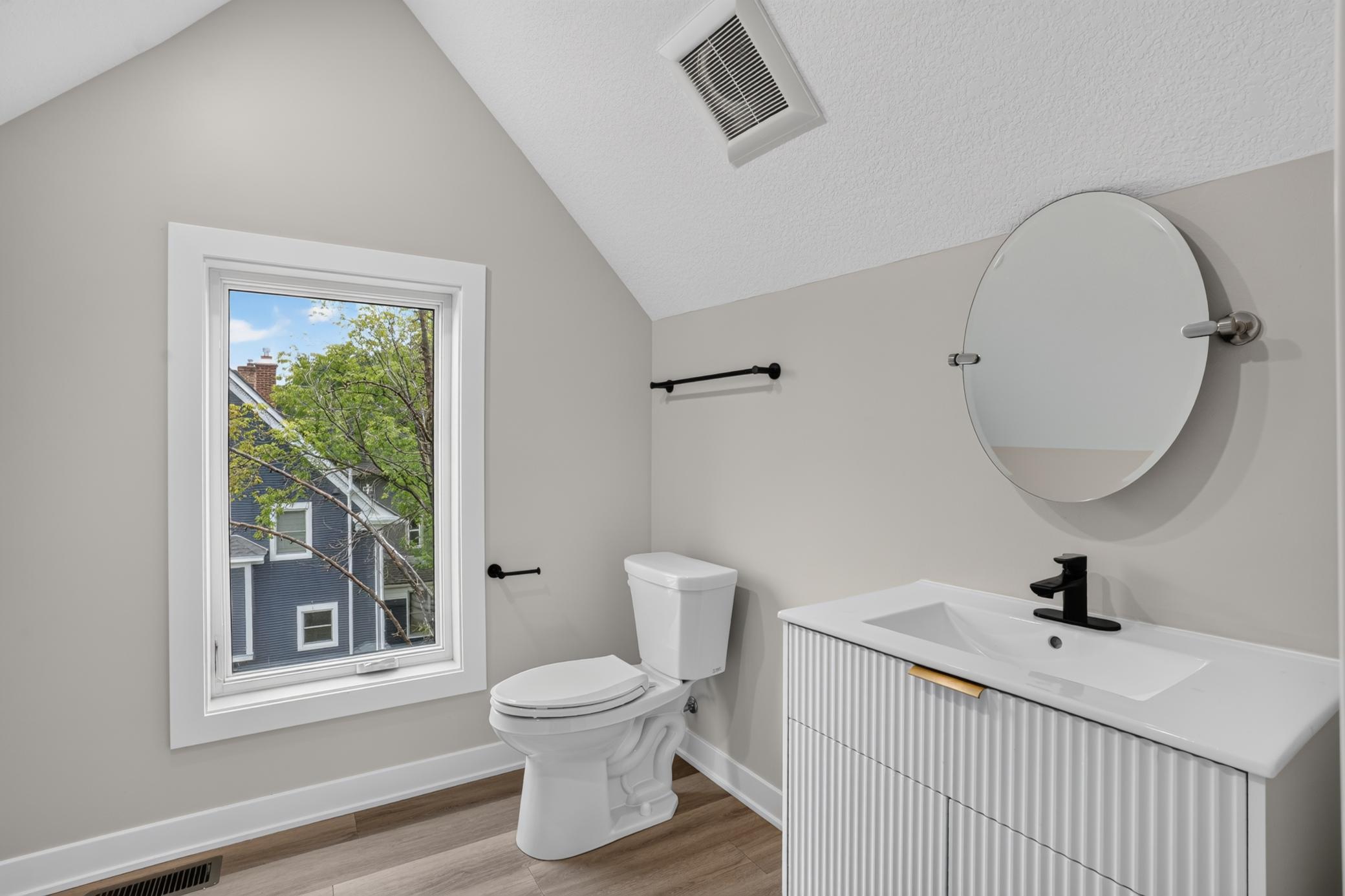 Bathroom with a fluted white vanity, round mirror, and window overlooking leafy trees.