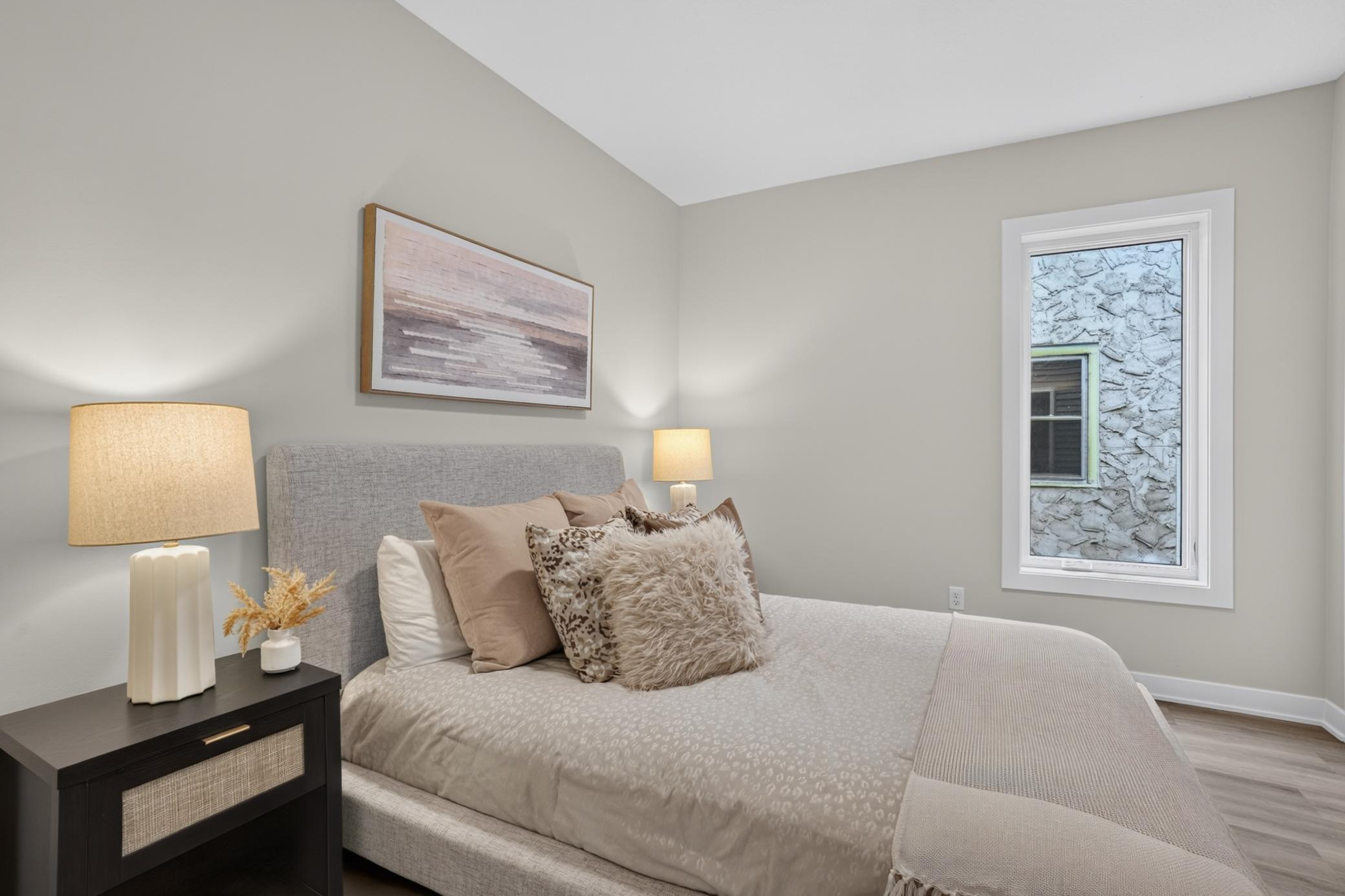 Furnished bedroom with a gray upholstered bed, neutral pillows, and a window beside the bed.