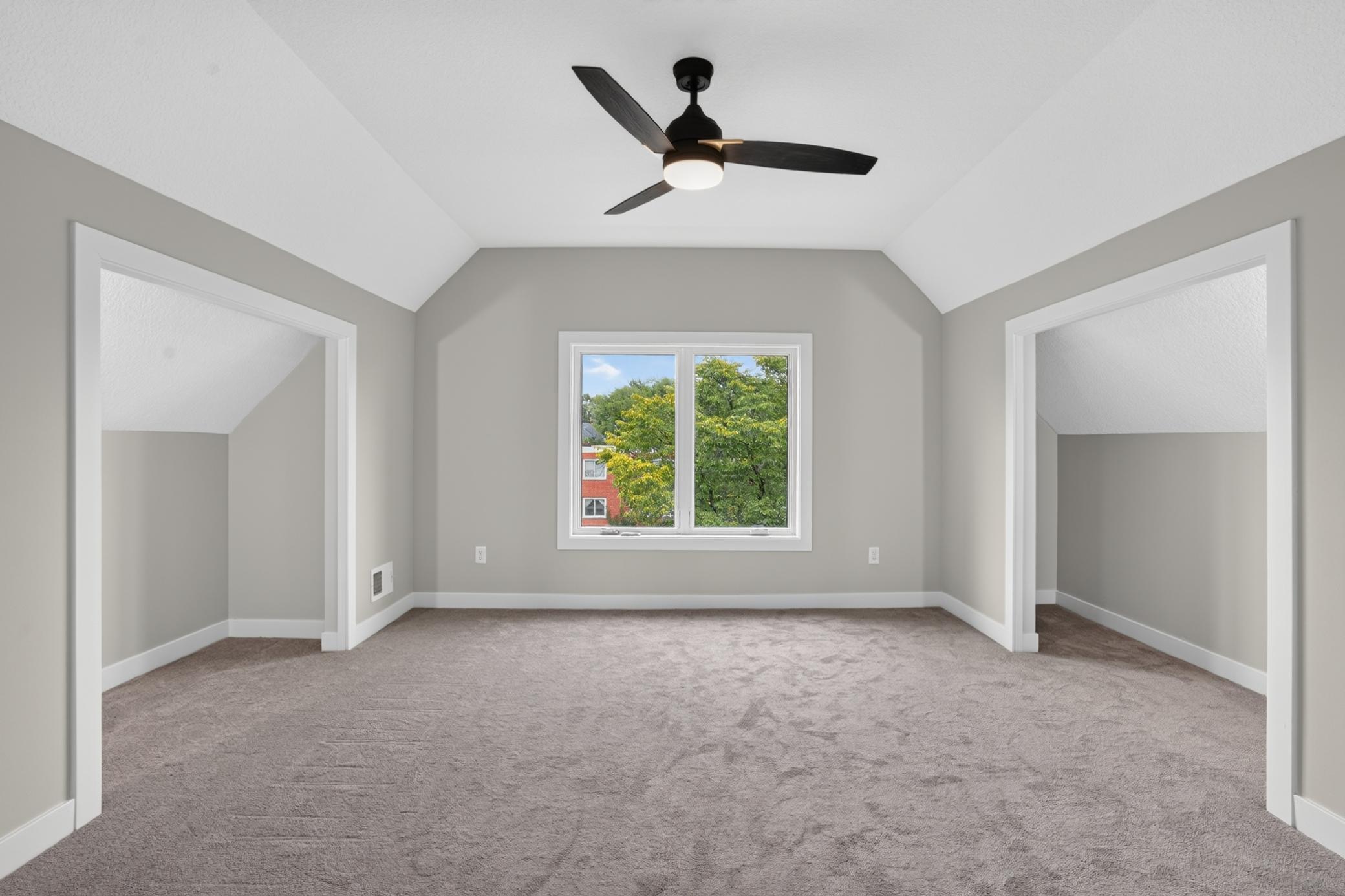 Empty carpeted room with sloped ceilings, a black ceiling fan, and a window overlooking trees.