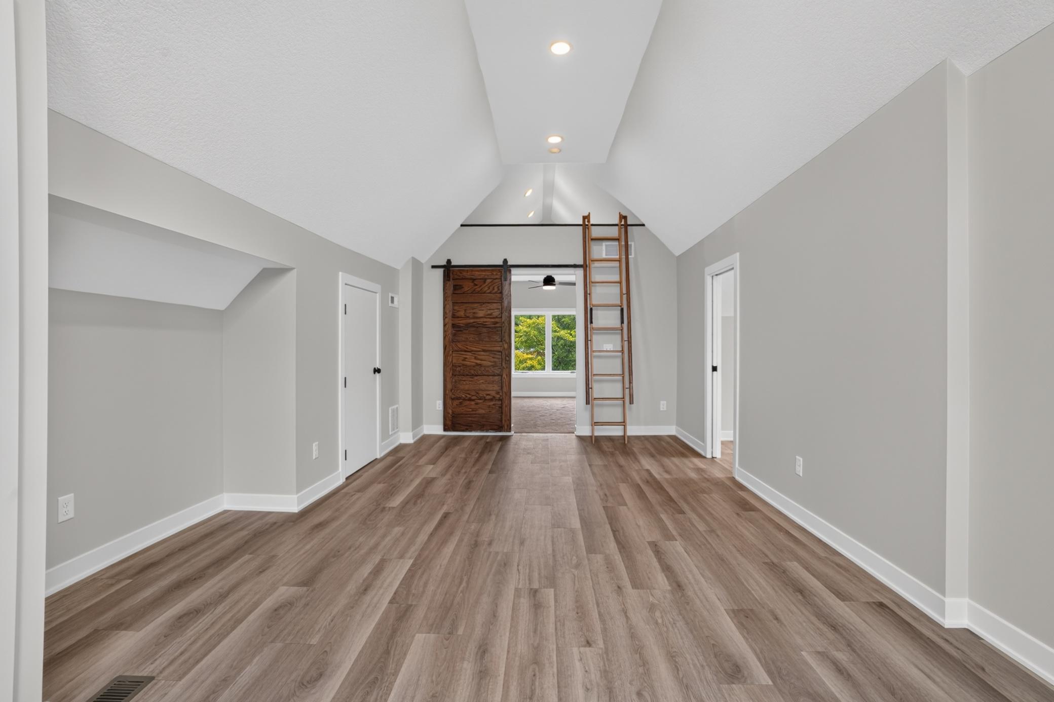 Vacant room with vaulted ceiling, wood-look floors, loft ladder, and sliding barn door.