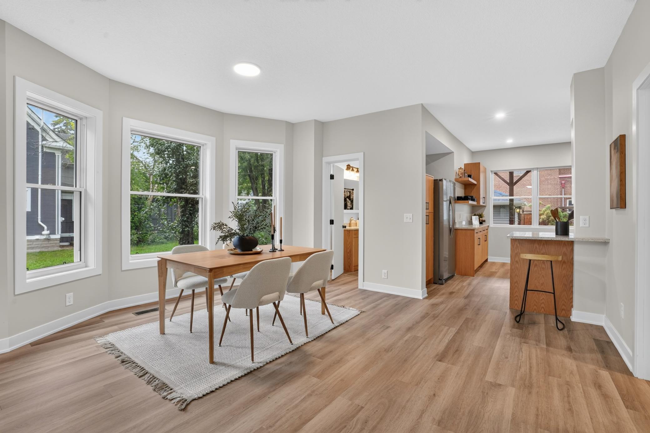 Open dining area beside a kitchen, with a wood table, large windows, and a wood-front island.