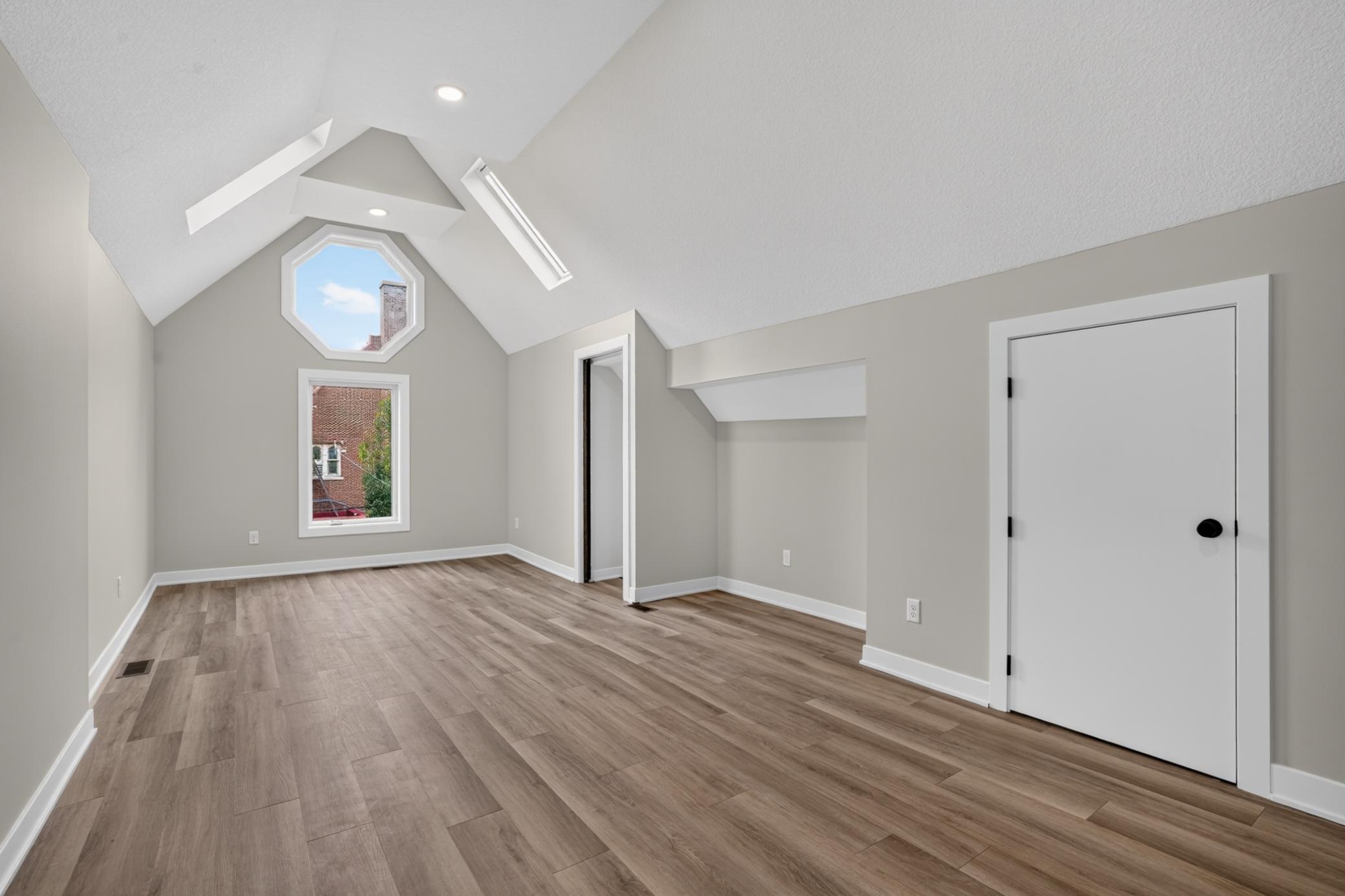 Empty attic room with vaulted ceiling, wood-look flooring, and an octagonal window above a rectangular one.