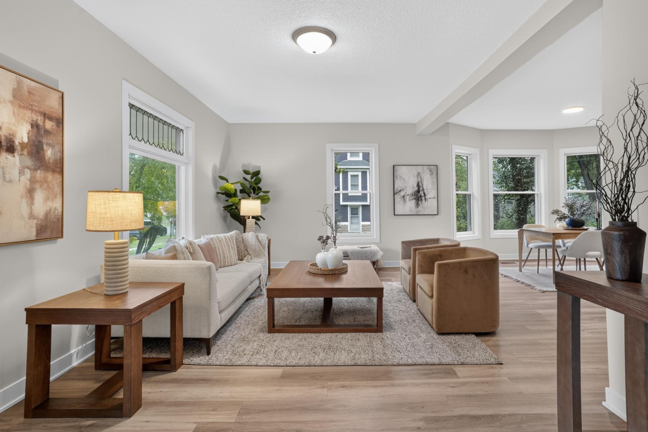 Staged living room with a cream sofa, brown chairs, and large windows beside the dining area.