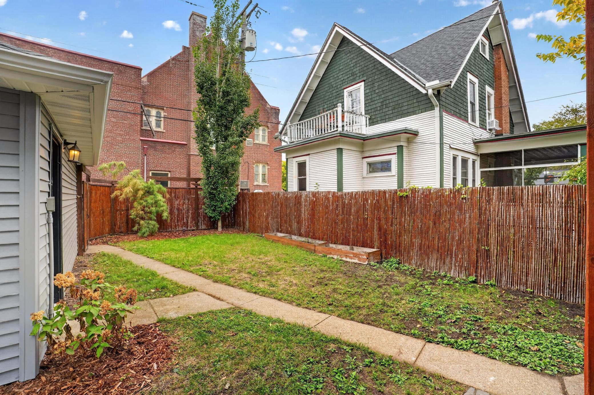 Fenced backyard with a small lawn, concrete path, and white house with dark green gables and balcony.