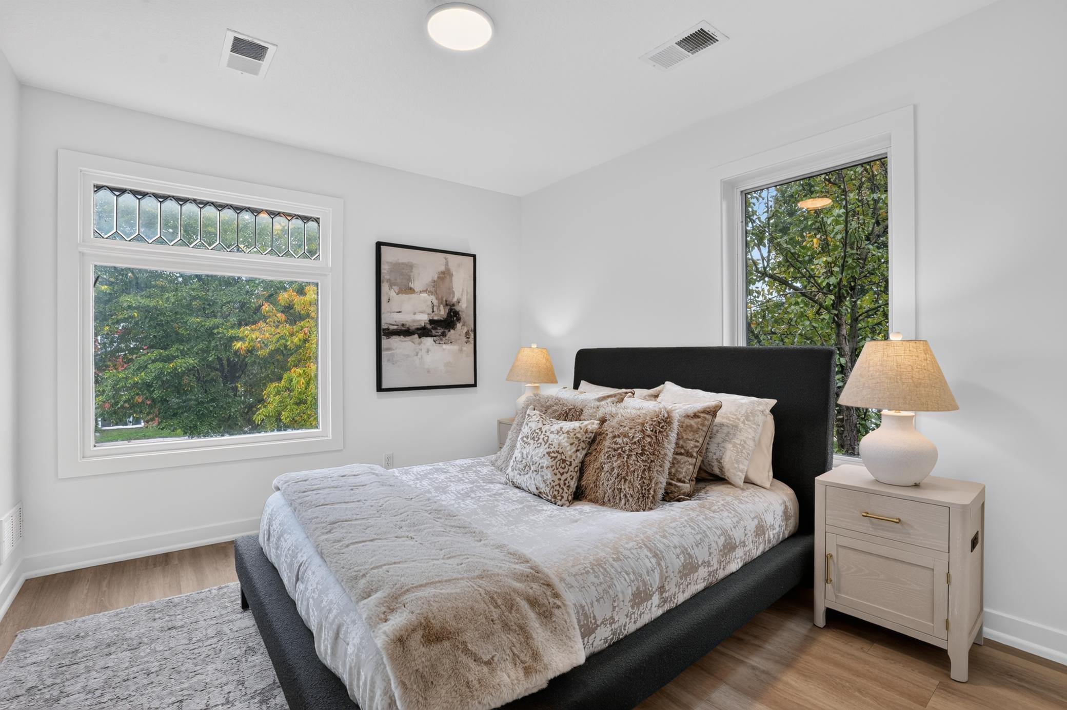 Bedroom with a dark upholstered bed, neutral layered bedding, and large windows overlooking leafy trees.