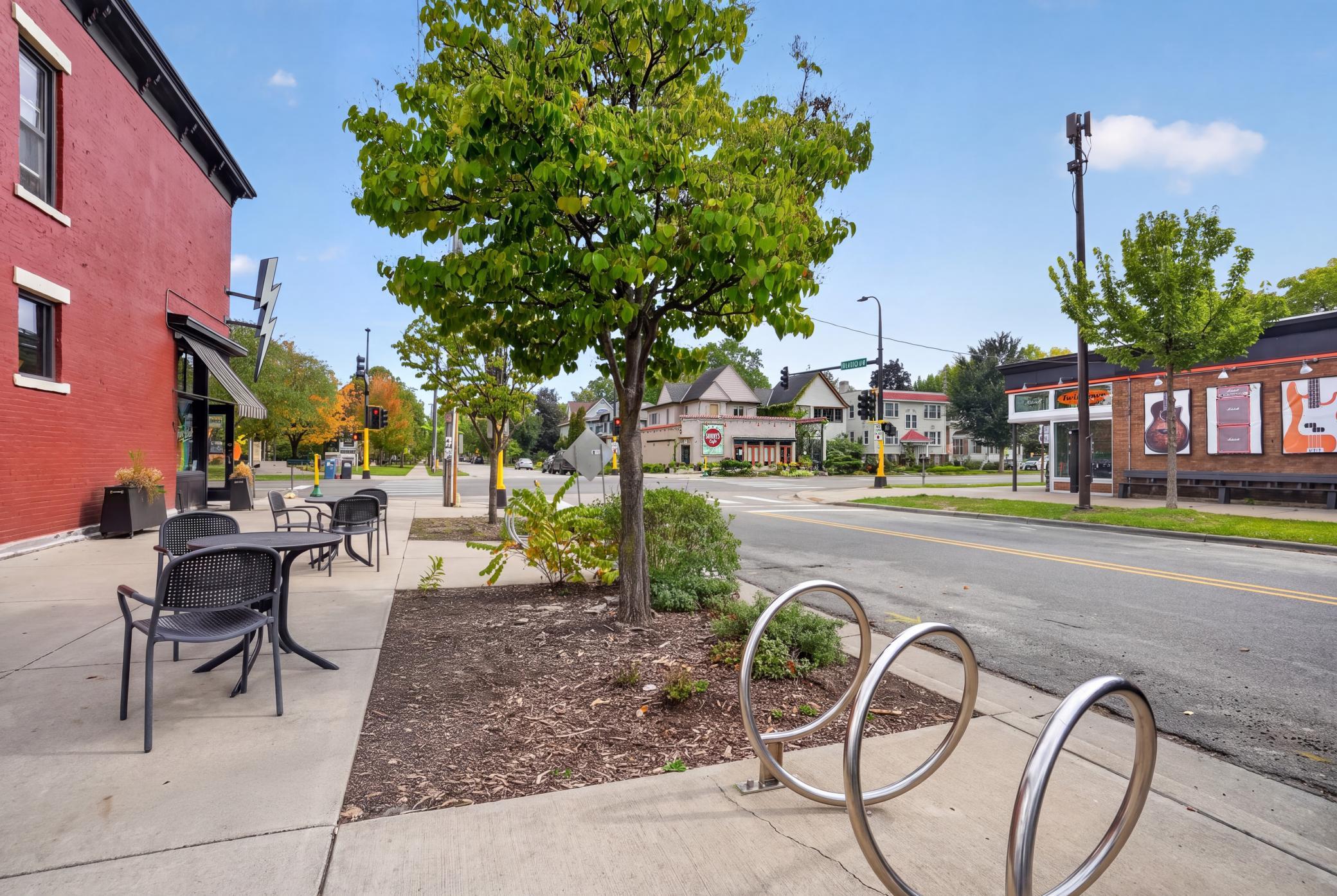 Neighborhood streetscape with sidewalk café seating, a leafy tree, and a bicycle rack at a shop-lined intersection.