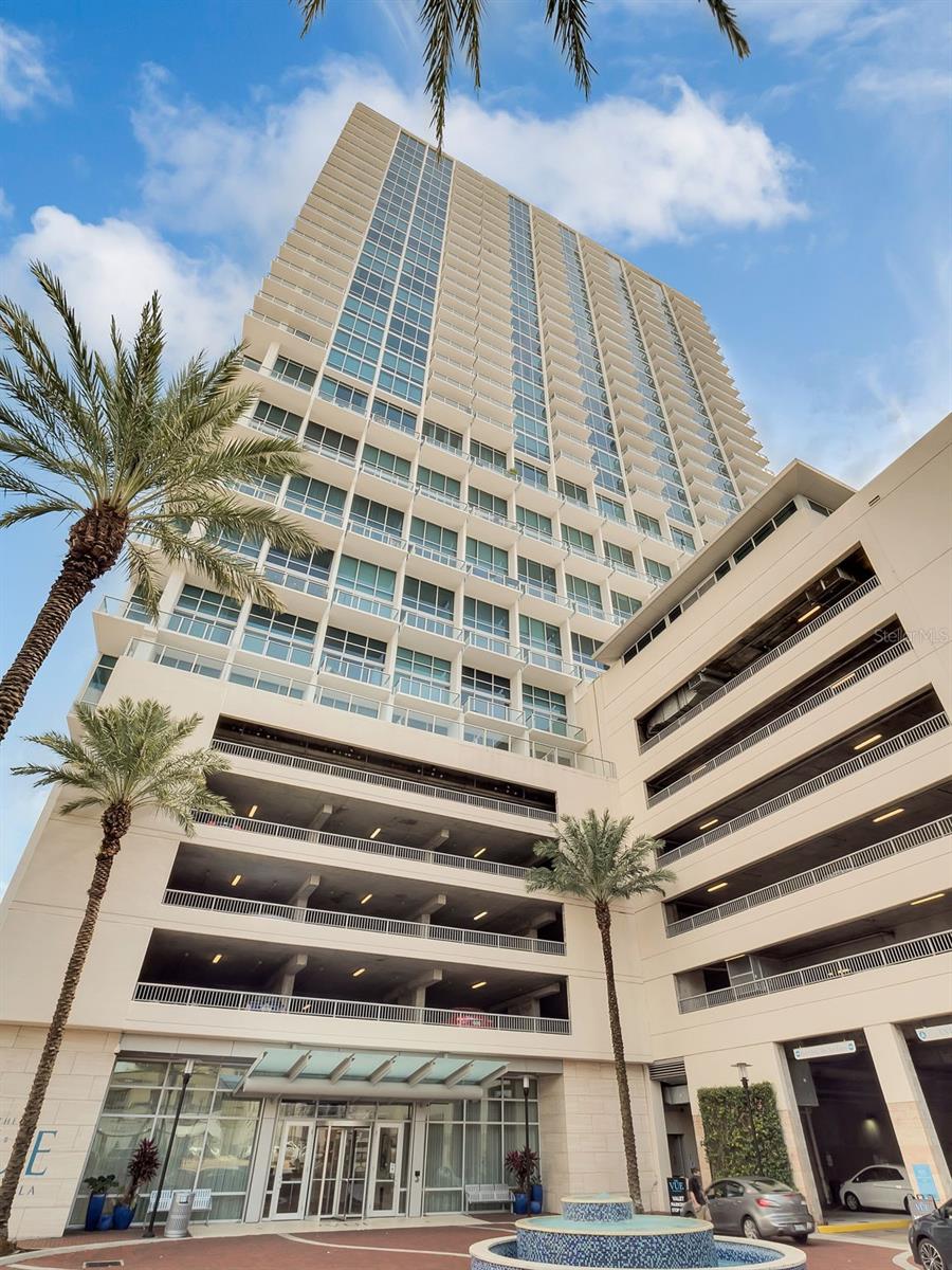 Modern high-rise condominium with glass balconies, an attached parking garage, palm trees, and a fountain.