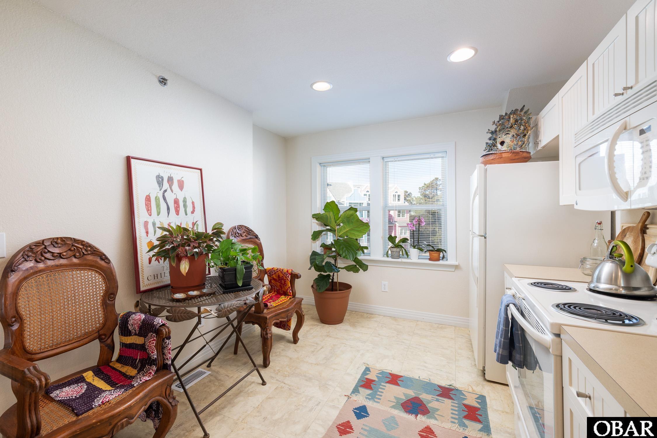 Kitchen with white appliances, carved wooden chairs, potted plants, and windows overlooking neighboring homes.