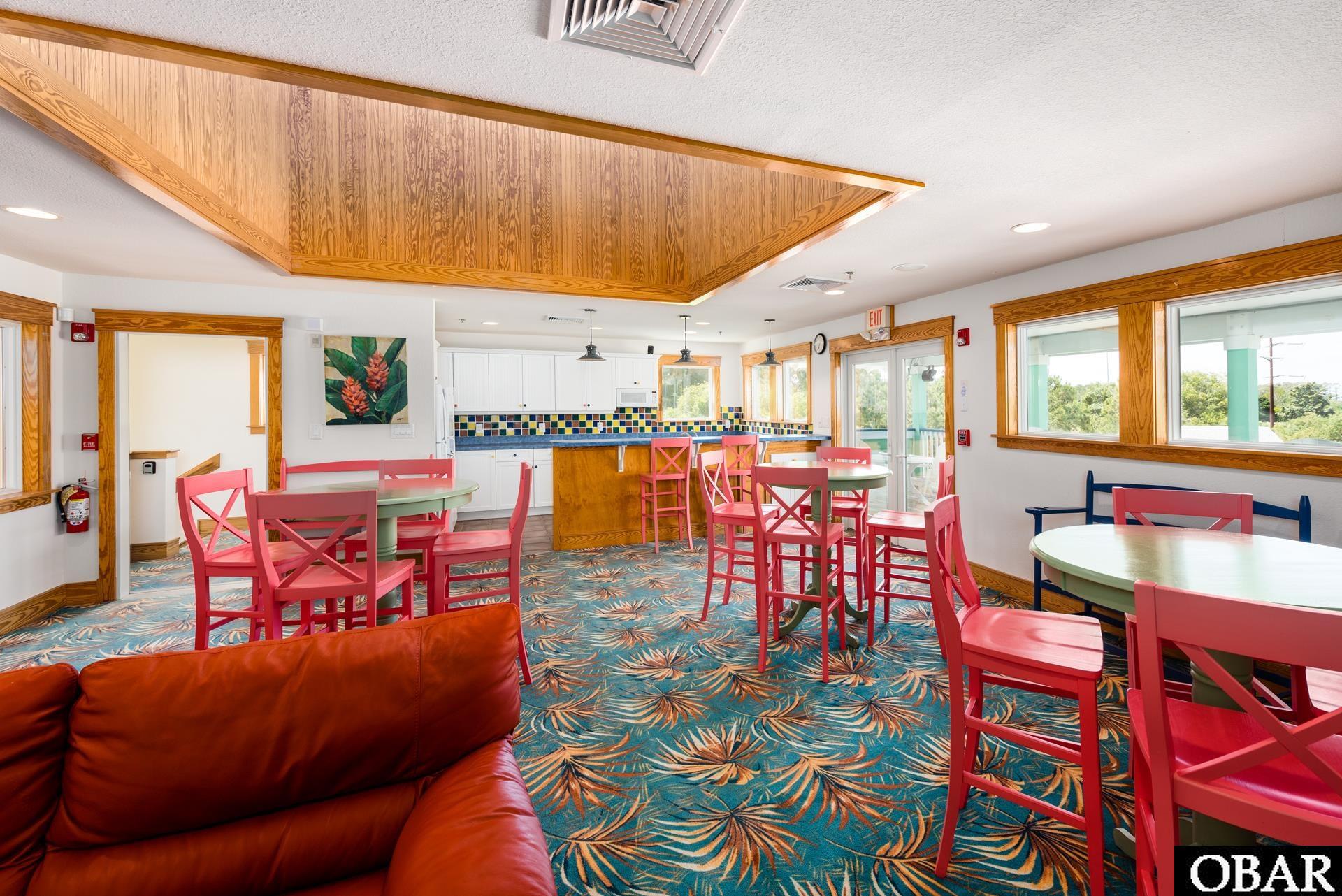 Furnished community lounge with a kitchen counter, red chairs, patterned carpet, and a wood-paneled ceiling.