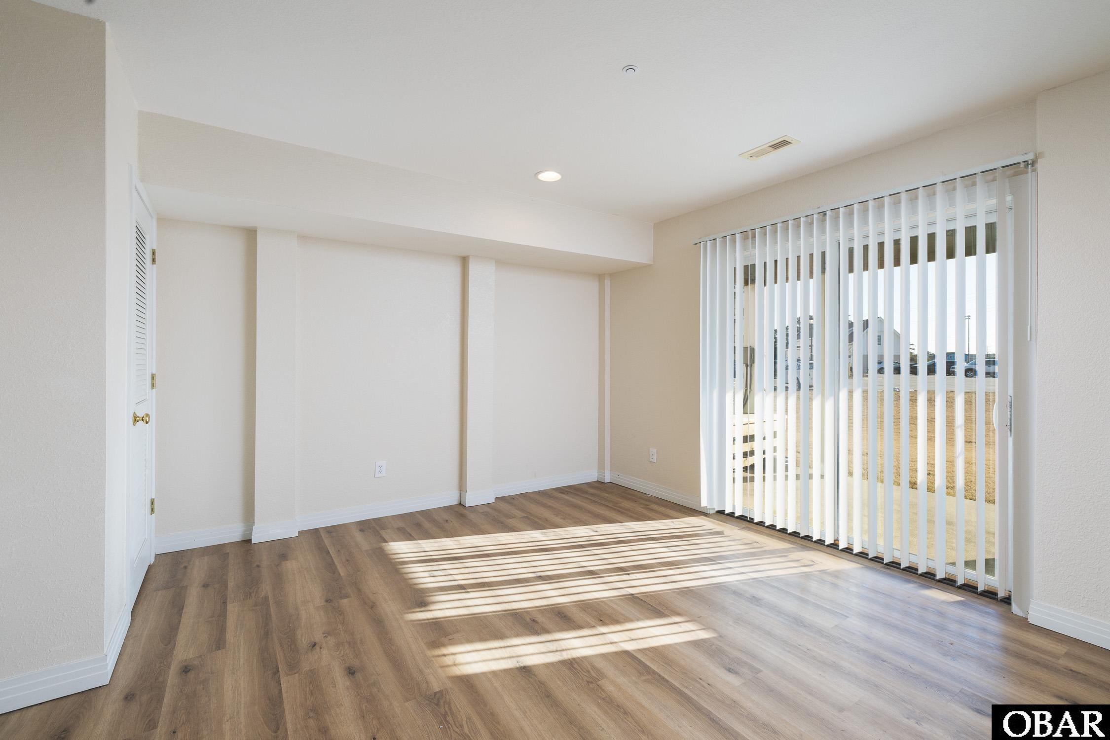 Empty living area with wood-look flooring and sunlight through vertical blinds.