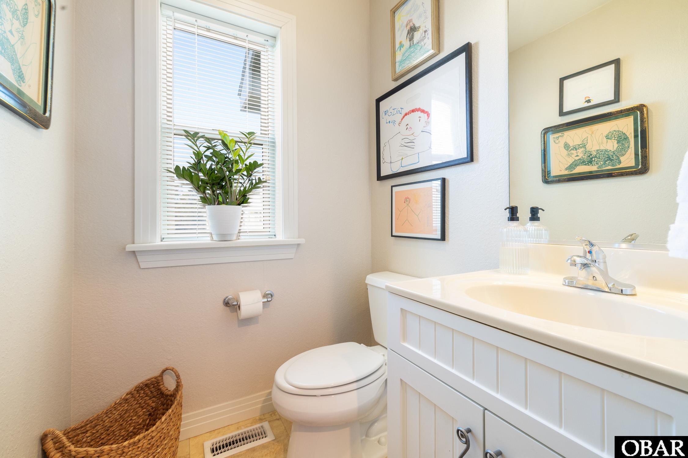 Bathroom with a white vanity and toilet, a window with blinds, and framed children’s artwork.