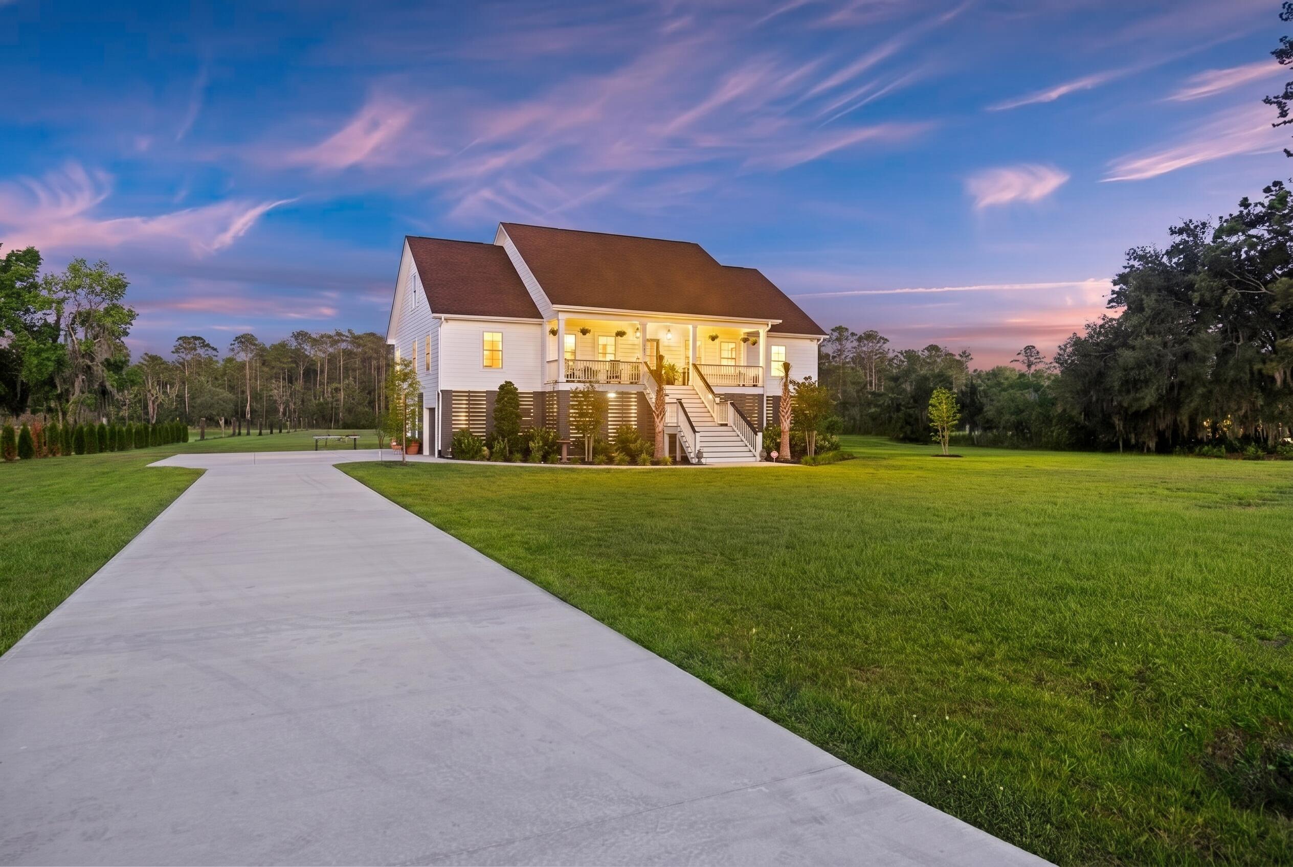 White raised house with a covered porch, long driveway, and broad lawn at dusk.