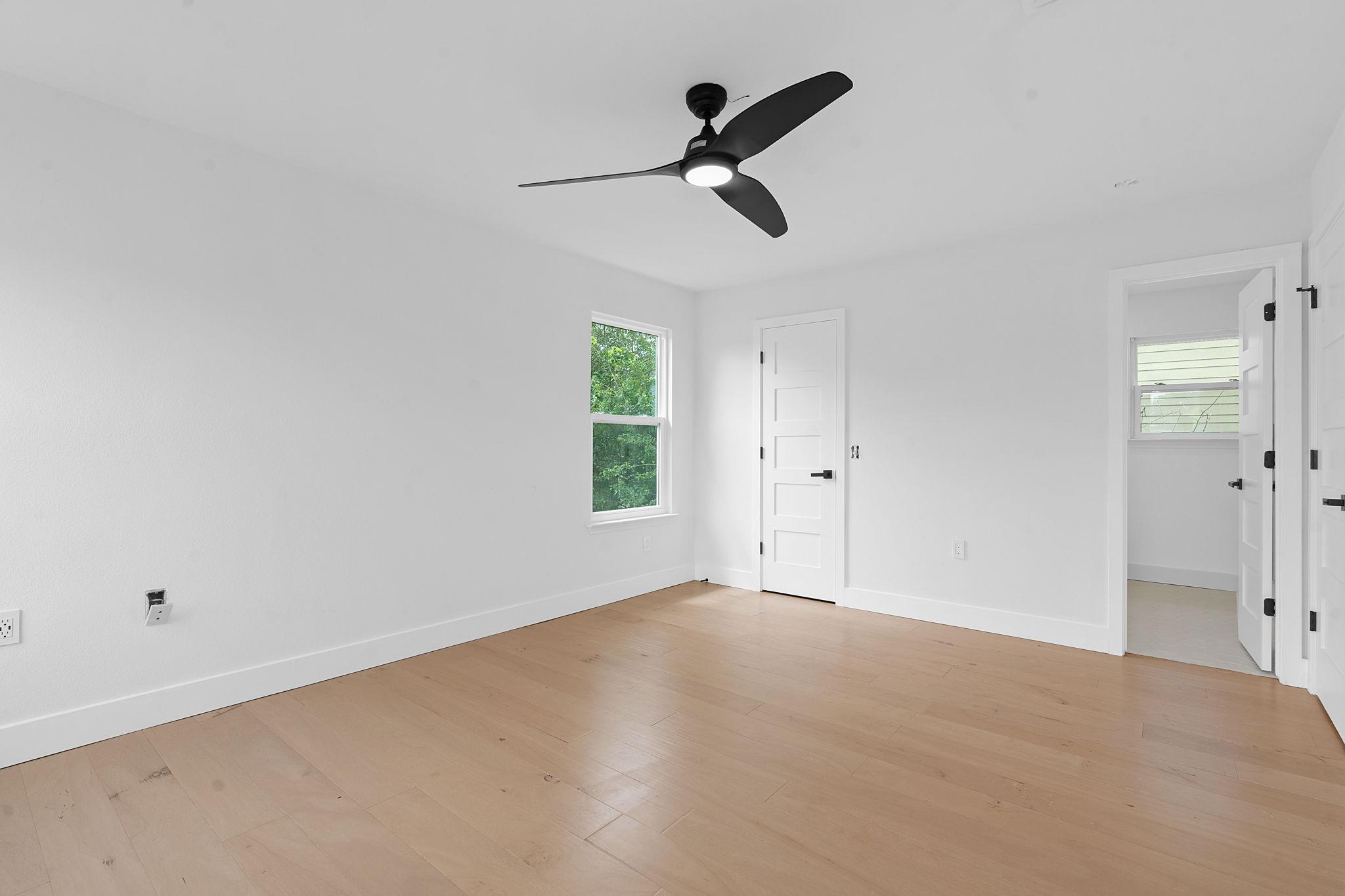Vacant bedroom with light wood floors, a black ceiling fan, and a window overlooking greenery.
