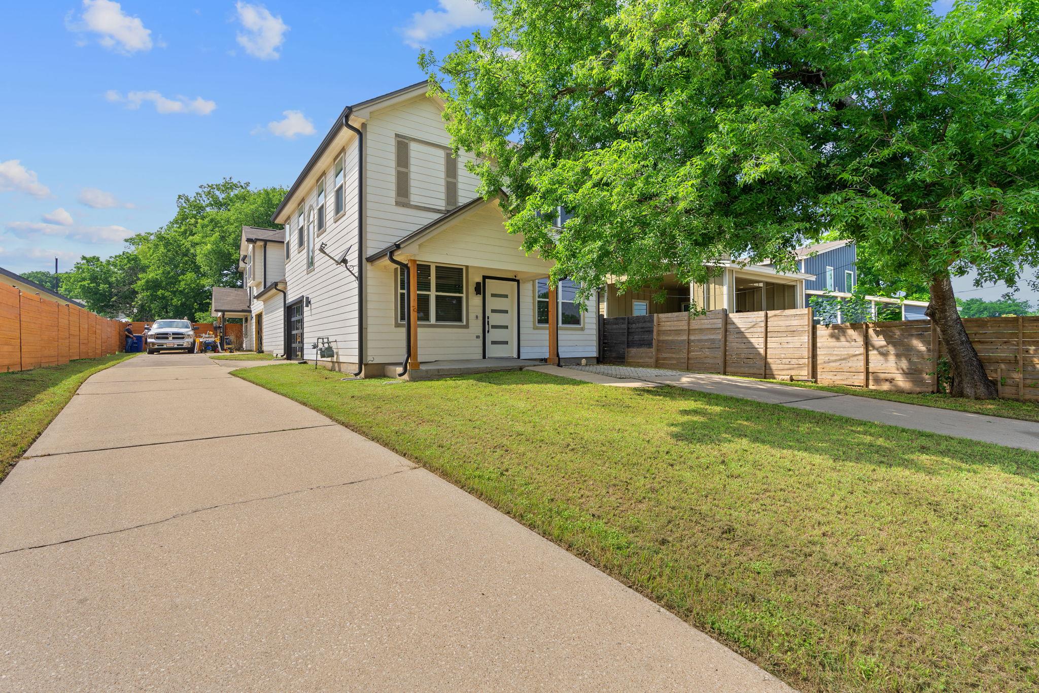 Two-story light-colored home with a covered entry, wide driveway, lawn, and wooden privacy fence.