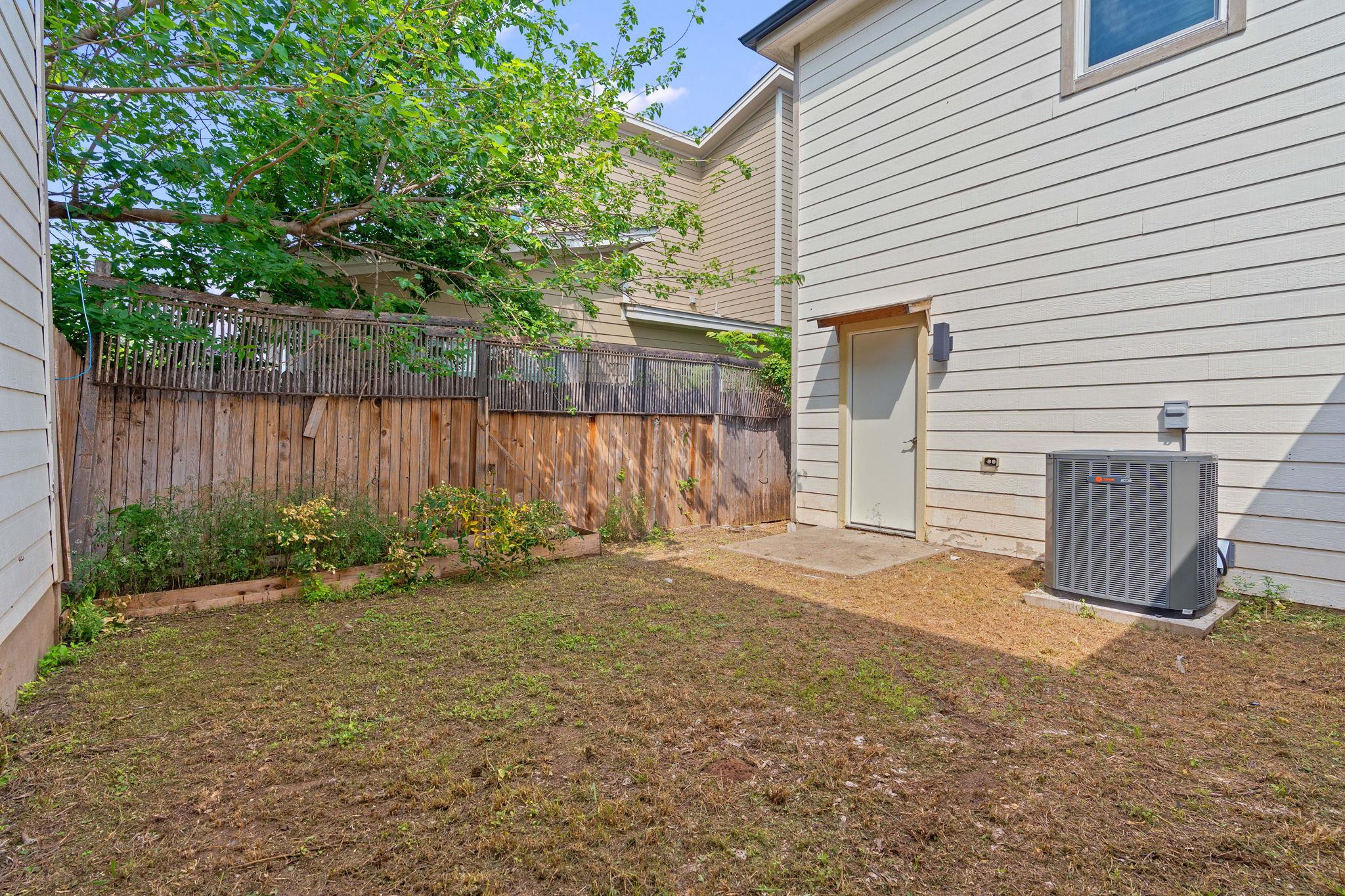 Fenced backyard with patchy lawn, raised planting beds, and a side door beside an air-conditioning unit.