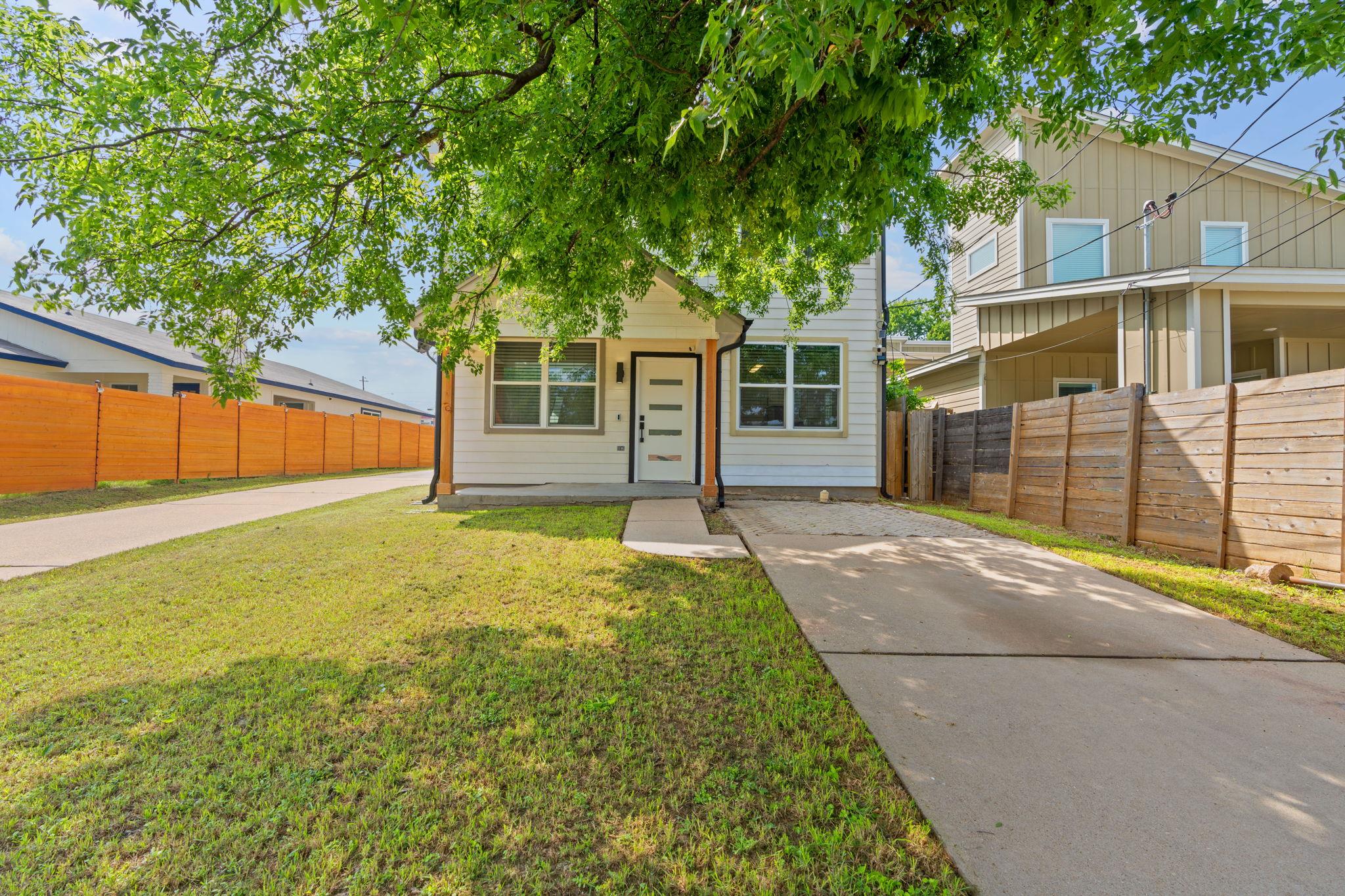 Small white-sided home with a concrete driveway, wooden fences, and a large leafy tree overhead.