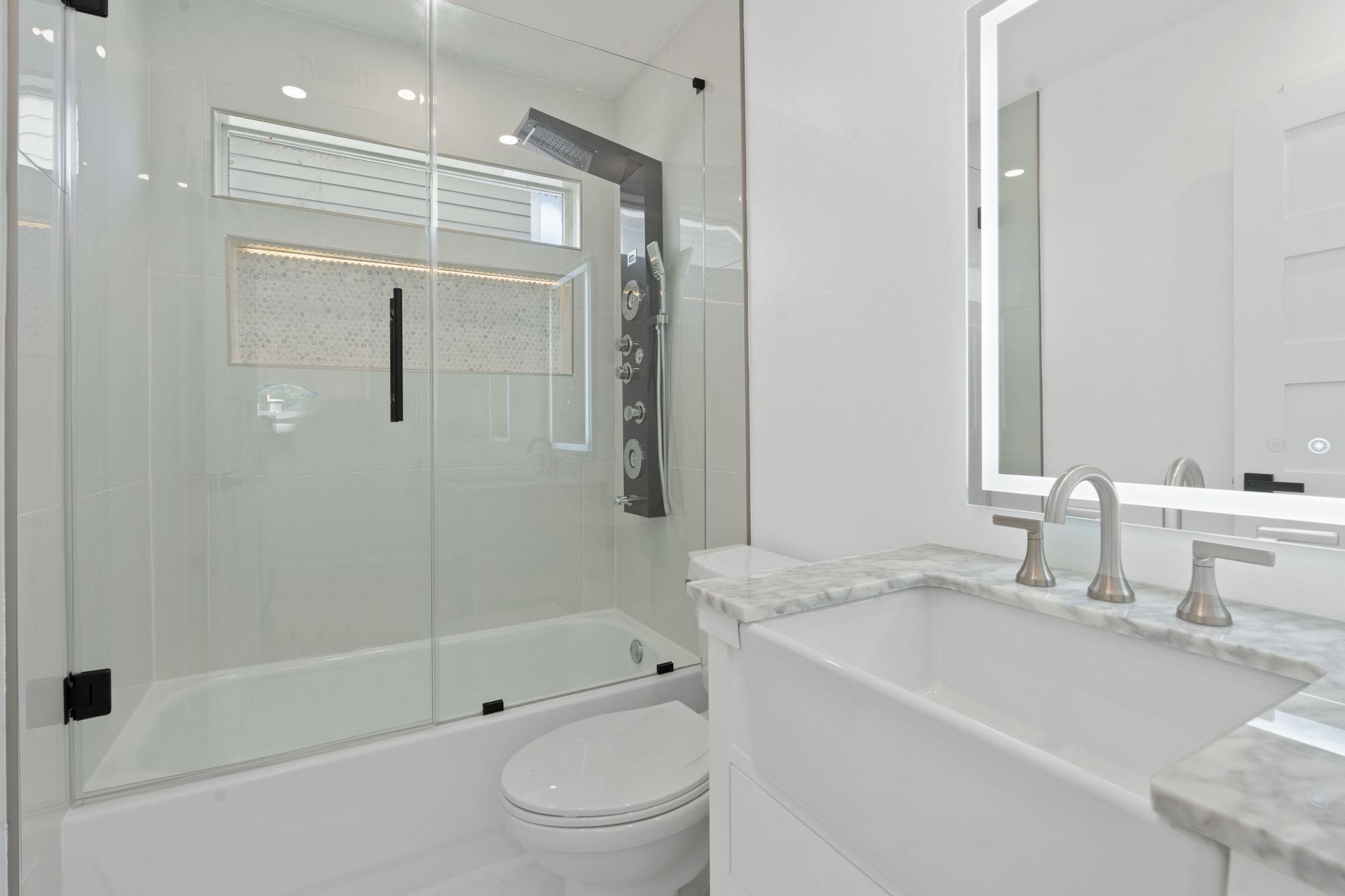 Bathroom with a glass-enclosed tub and shower, multi-jet shower panel, marble vanity, and illuminated mirror.