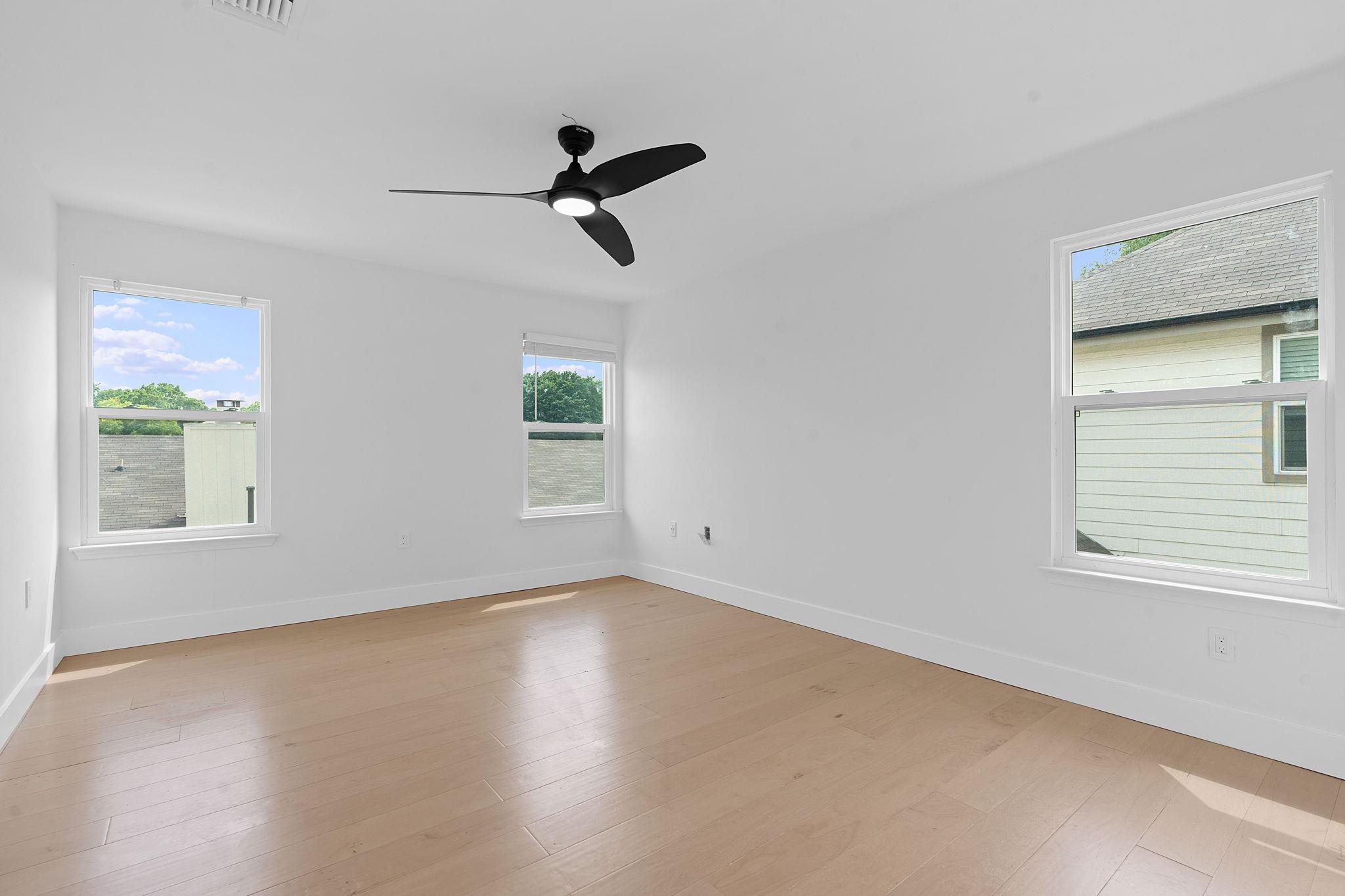 Vacant room with light wood floors, three windows, and a black ceiling fan.