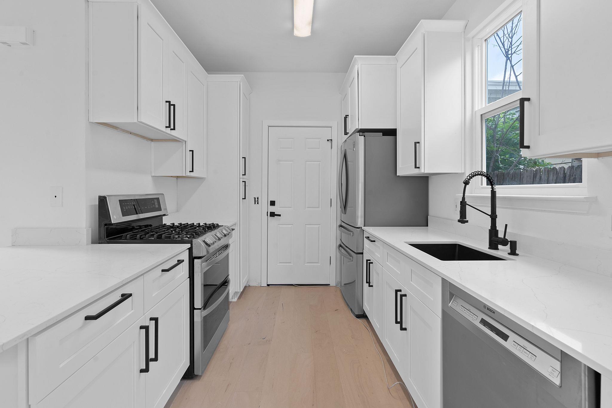 Renovated white kitchen with quartz counters, stainless appliances, a gas range, and a black sink beneath a window.
