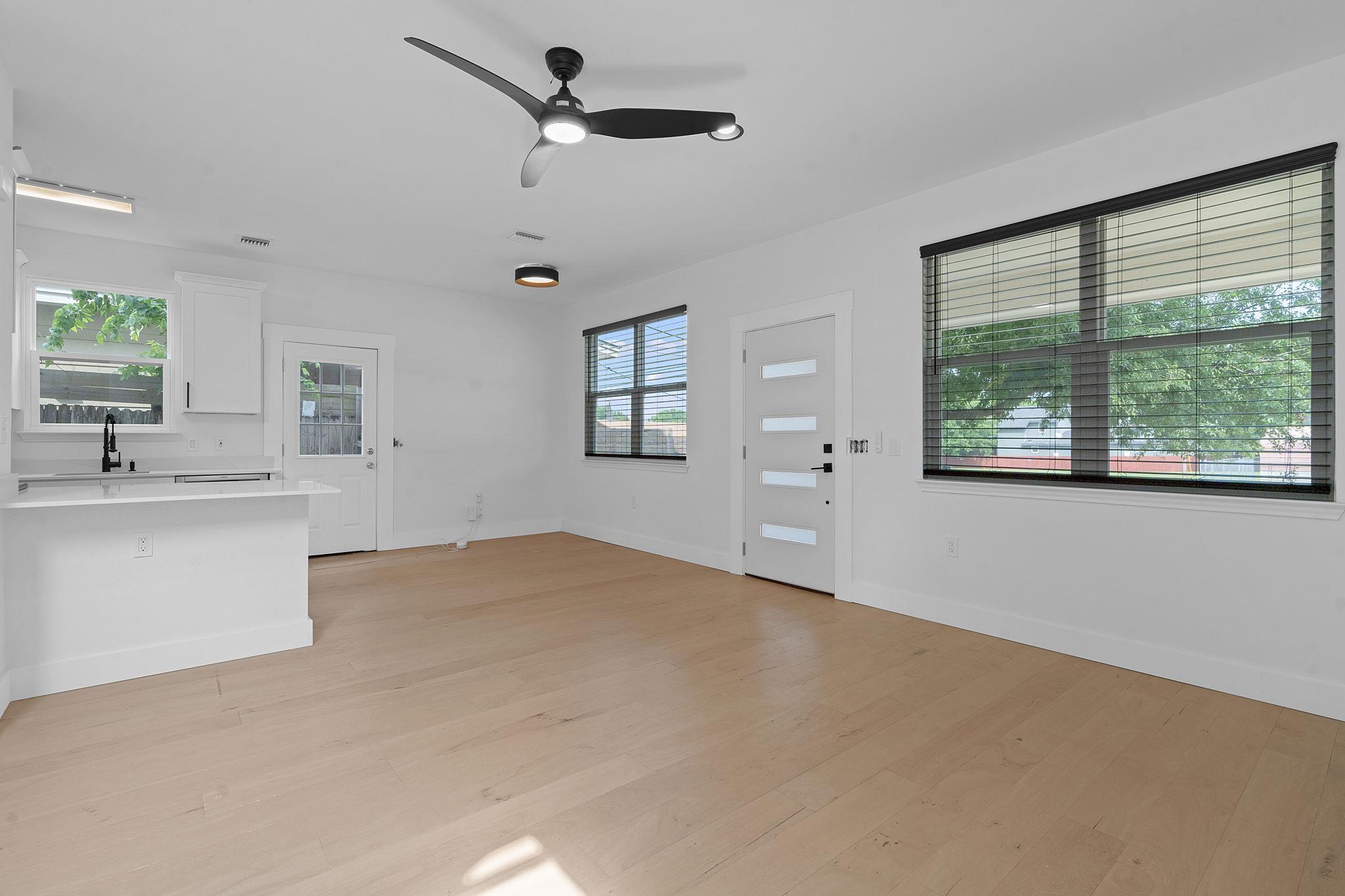 Vacant living area with pale wood floors, kitchen peninsula, ceiling fan, and large windows with blinds.