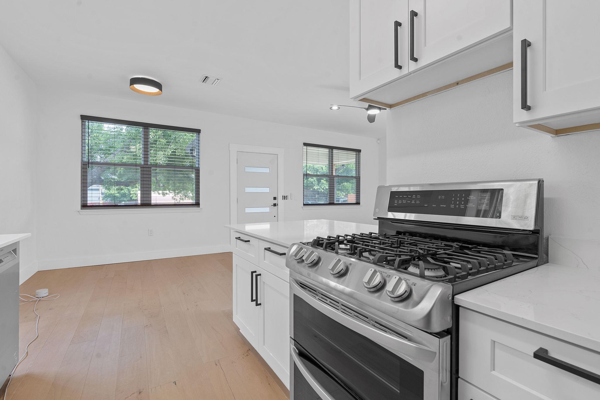 Modern kitchen with a large stainless steel gas range, white cabinets, pale wood floors, and two windows.