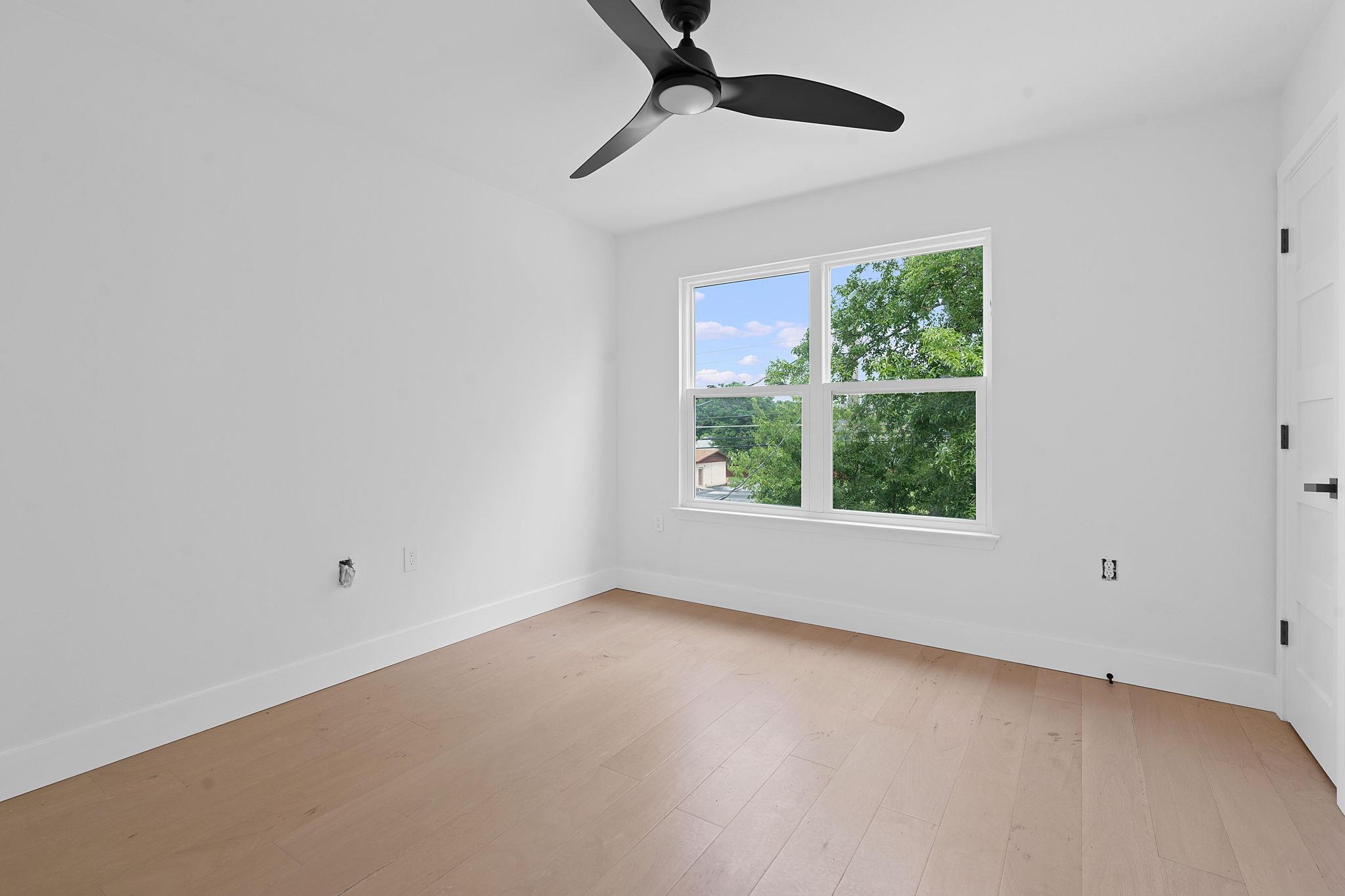 Empty bright room with pale wood floors, a black ceiling fan, and a large window.