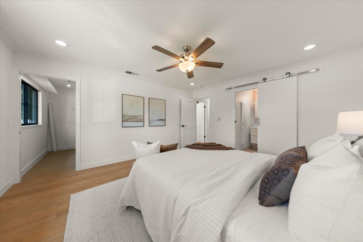 Staged bedroom with a white bed, hardwood floors, ceiling fan, and sliding barn door.
