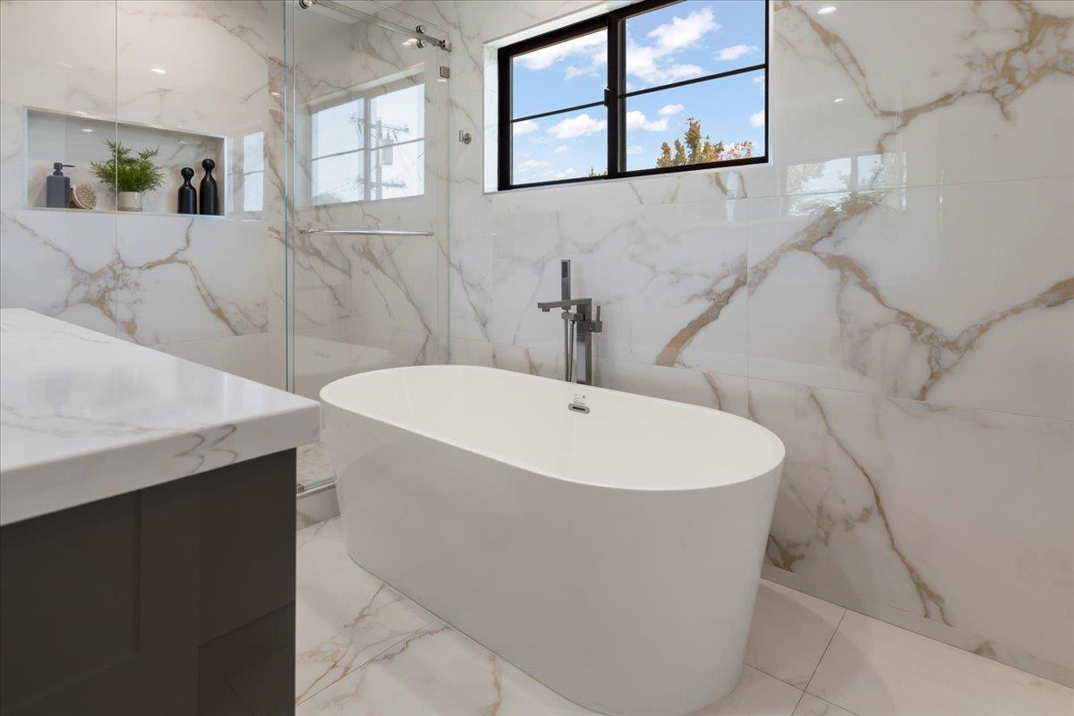 Bathroom with a freestanding white tub, marble-look tile, glass shower, and black-framed window.