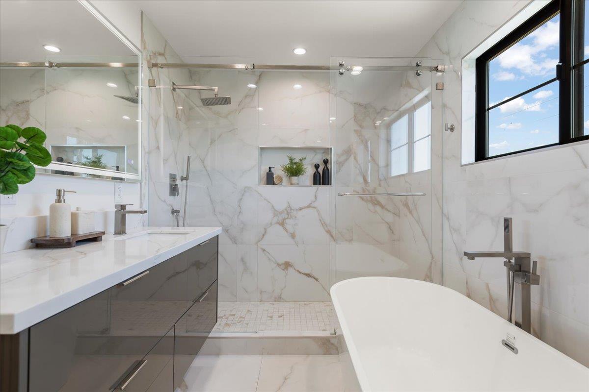Bathroom with a freestanding tub, glass shower, marble-look tile and black-framed window.