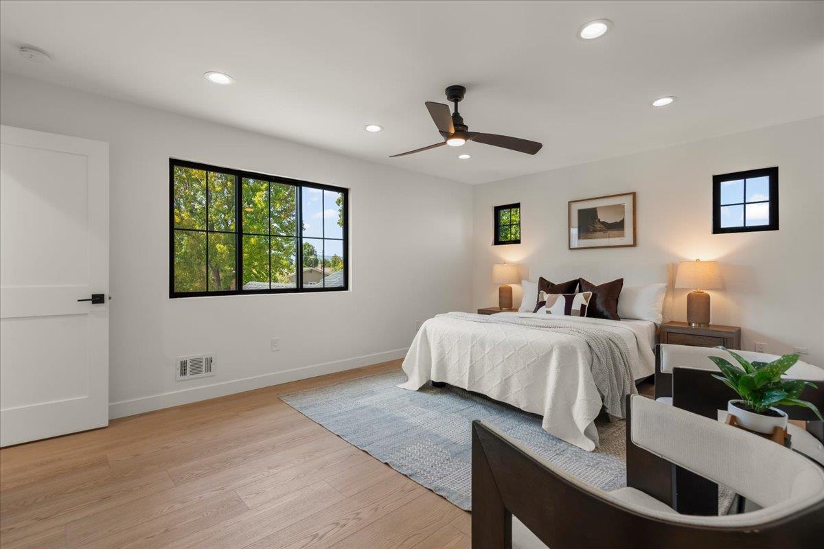 Staged bedroom with a white bed, ceiling fan, and large black-framed windows overlooking leafy trees.