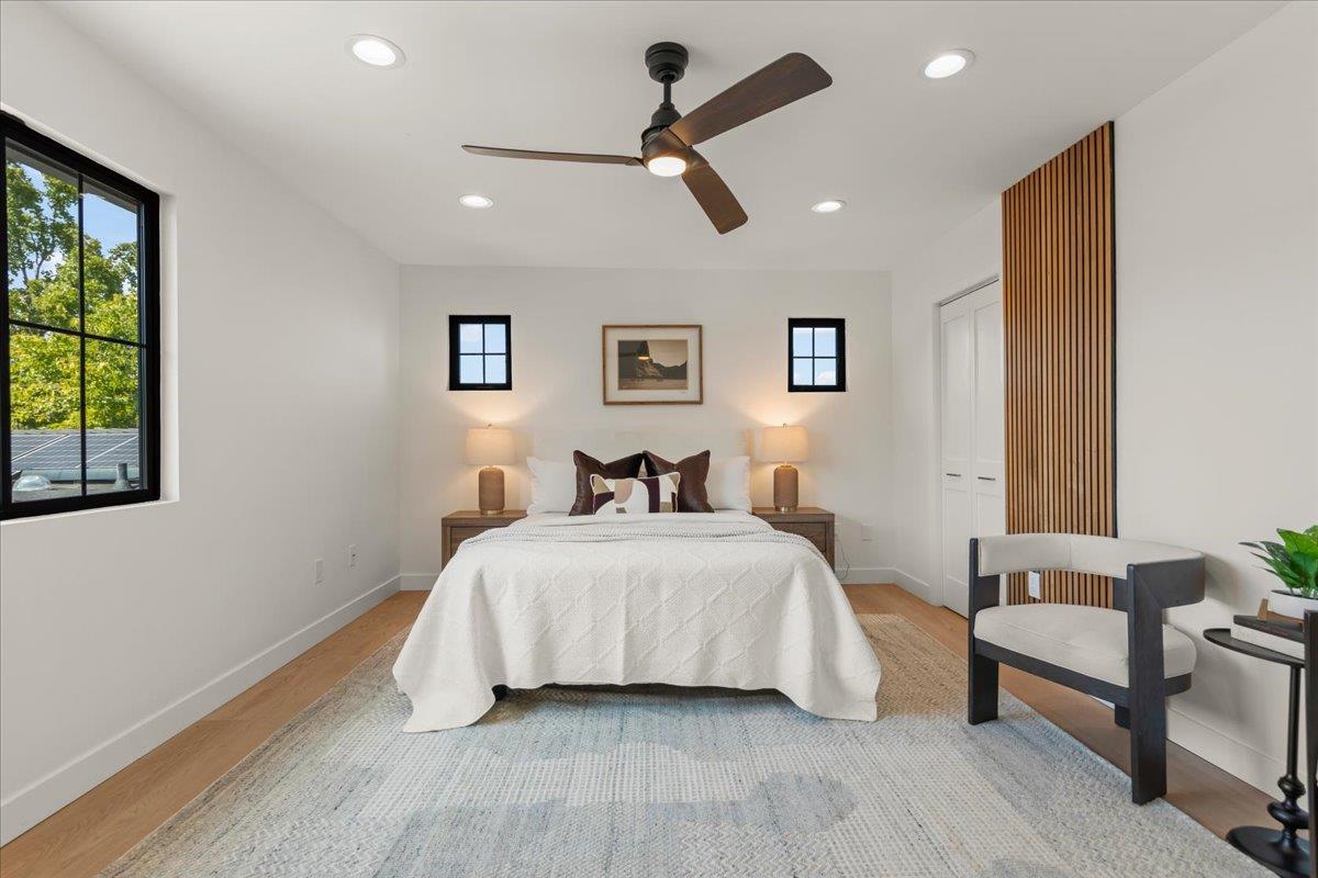 Staged bedroom with a white bed, large black-framed window, wood-slat accent, and dark ceiling fan.