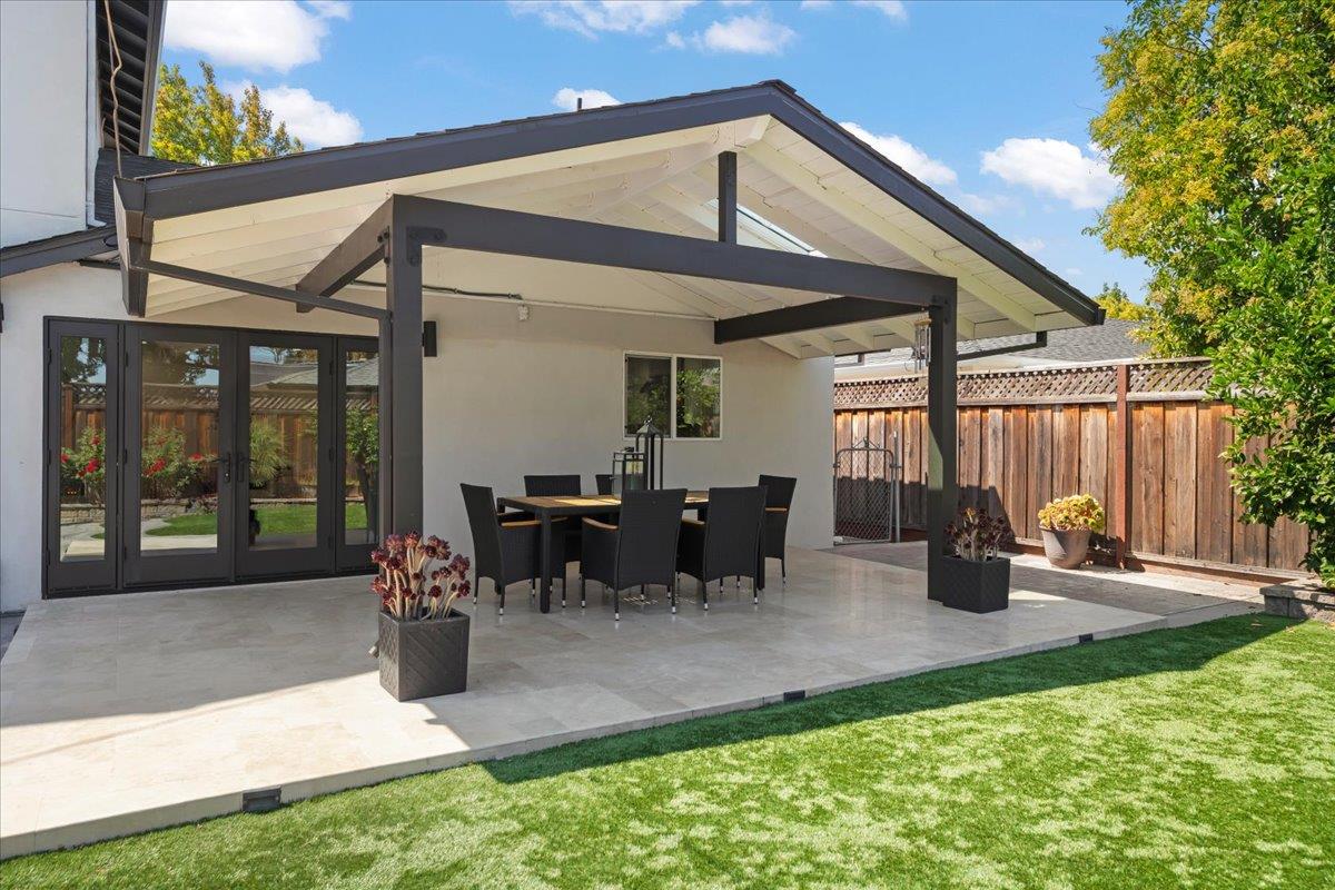 Covered backyard patio with dark beams, outdoor dining set, pale stone paving, and lawn.