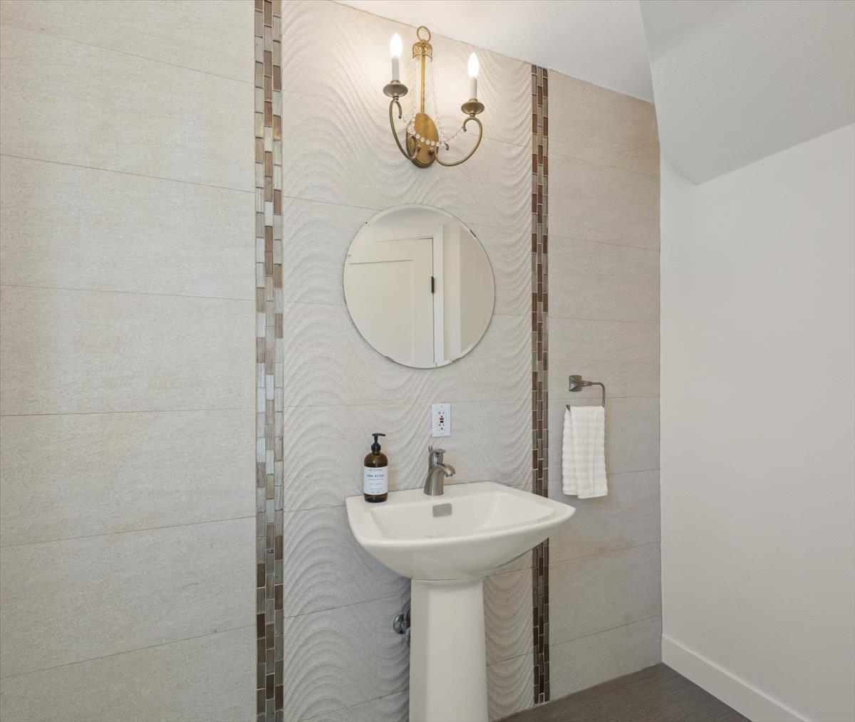 Bathroom pedestal sink beneath a round mirror and brass sconce, set against textured tile.